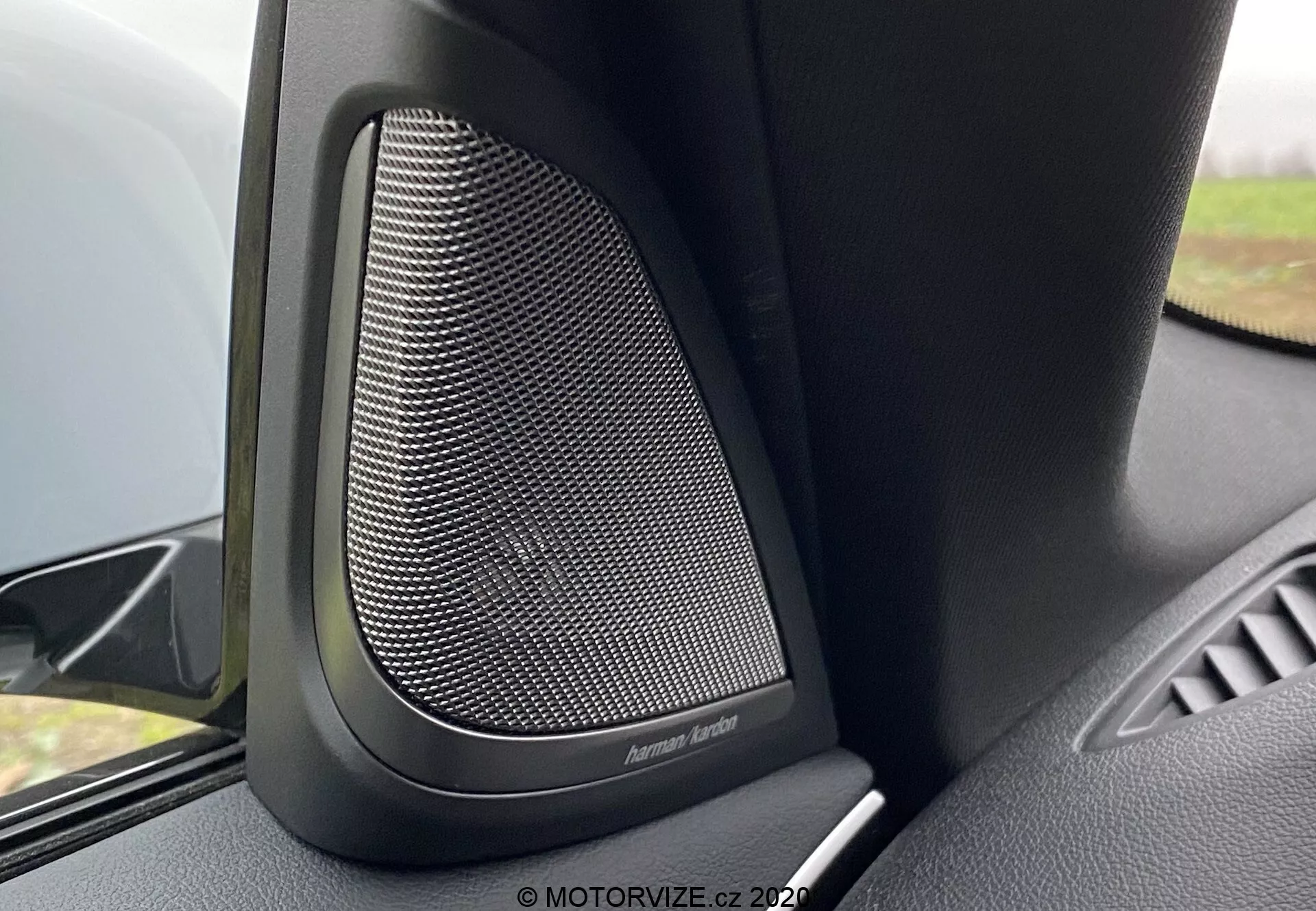 Zbliżenie na wnętrze BMW 5er-Reihe 2020 (BMW 520i Touring Steptronic) skupiające się na zaawansowanym głośniku drzwiowym Harman Kardon. Zdjęcie ukazuje delikatny siatkowy wzór metalowej kratki osłaniającej głośnik, otoczonej czarnym plastikiem lub miękkim materiałem tłumiącym, aby redukować niechciane wibracje i zapewniać najwyższą jakość dźwięku. Kąt kamery pokazuje górną część panelu drzwi, z wyeksponowanym głośnikiem i niewielką częścią podłokietnika drzwi. Delikatne rozmazanie tła podkreśla ten wysokiej klasy system audio w luksusowym pojeździe.