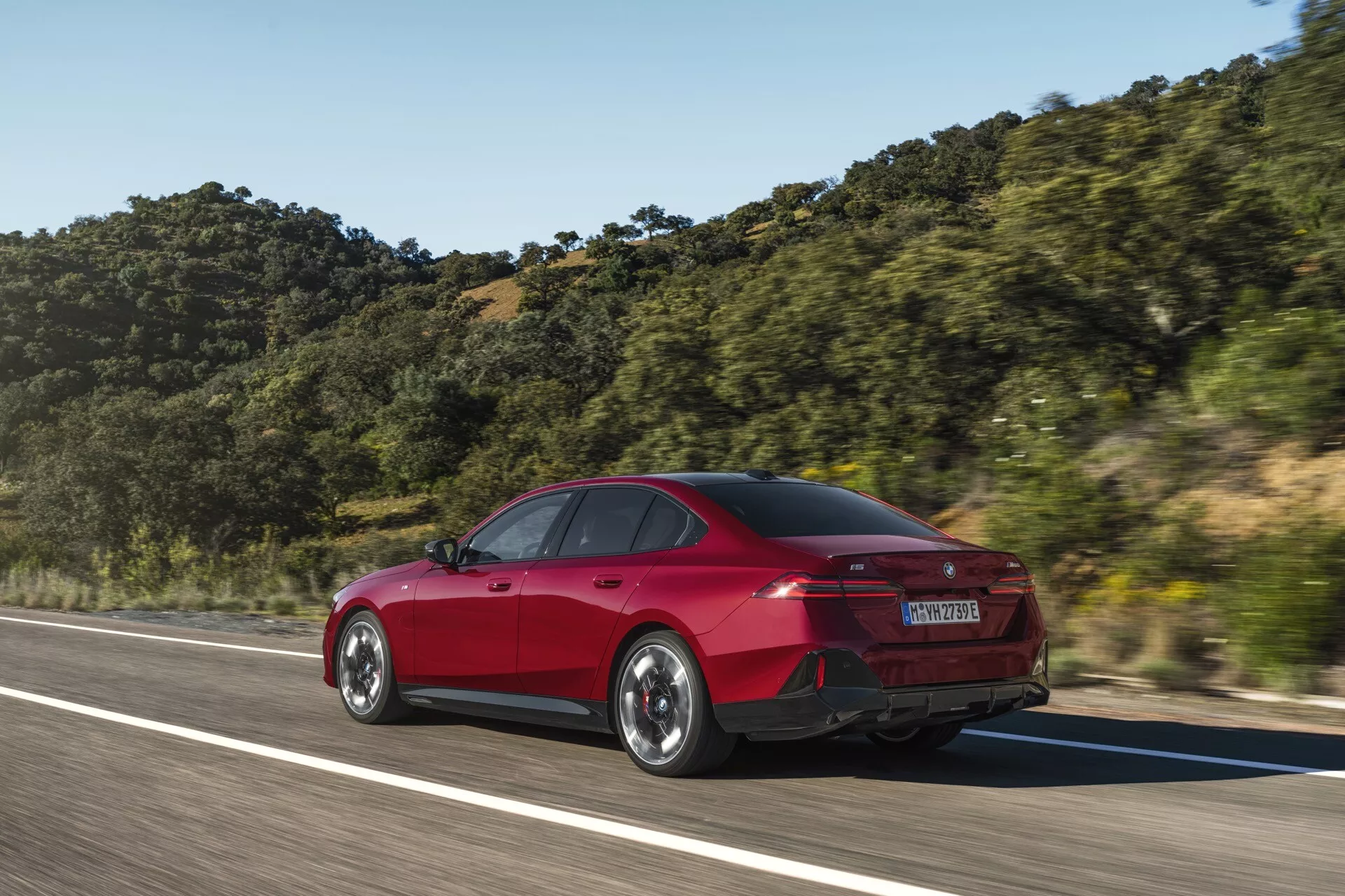 Czerwony sedan BMW serii 5 (G60-G61) z czterema drzwiami w ruchu, ujęty pod kątem trzy czwarte od tyłu, podkreślający tylną i boczną stylizację jak kształt bagażnika, zderzak tylny, tylne lampy LED i linie aerodynamiczne na tle górskim pod jasnym niebem