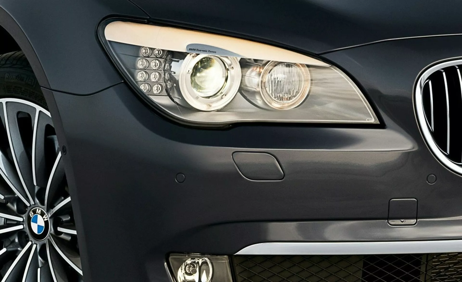 Zbliżenie przedniej części bocznej BMW serii 7 z 2005 roku z emblematem marki, nowoczesnym zespołem reflektorów z soczewką projektora i diodami LED do jazdy dziennej, częściowym widokiem na atrapę chłodnicy nerkową i wieloramienne koło ze stopu lekkiego.