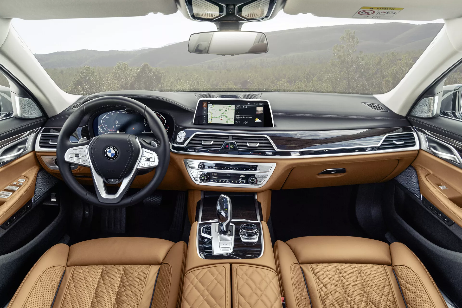Obraz przedstawia wnętrze BMW Serii 7 z 2019 roku (lift G11/G12) z perspektywy tylnego siedzenia, podkreślając szczegółowy i luksusowy design przedniego kokpitu. Z nowoczesną kierownicą z przyciskami multifunkcyjnymi i wyraźnym emblematem BMW, elegancka deska rozdzielcza mieści centralny dotykowy wyświetlacz infotainment. Konsola środkowa zawiera automatyczną dźwignię zmiany biegów, touchpad i różne systemy kontroli dynamicznej, uzupełnione beżowymi, pikowanymi skórzanymi siedzeniami sygnalizującymi poziom wyposażenia premium. Panele drzwi odbijają ten luksus z beżową skórą, drewnianymi wykończeniami i oświetleniem ambient, sugerując, że zaawansowane funkcje technologiczne i komfortowe są zintegrowane w całym pojeździe, w tym dach panoramiczny zapewniający dodatkowe naturalne światło.