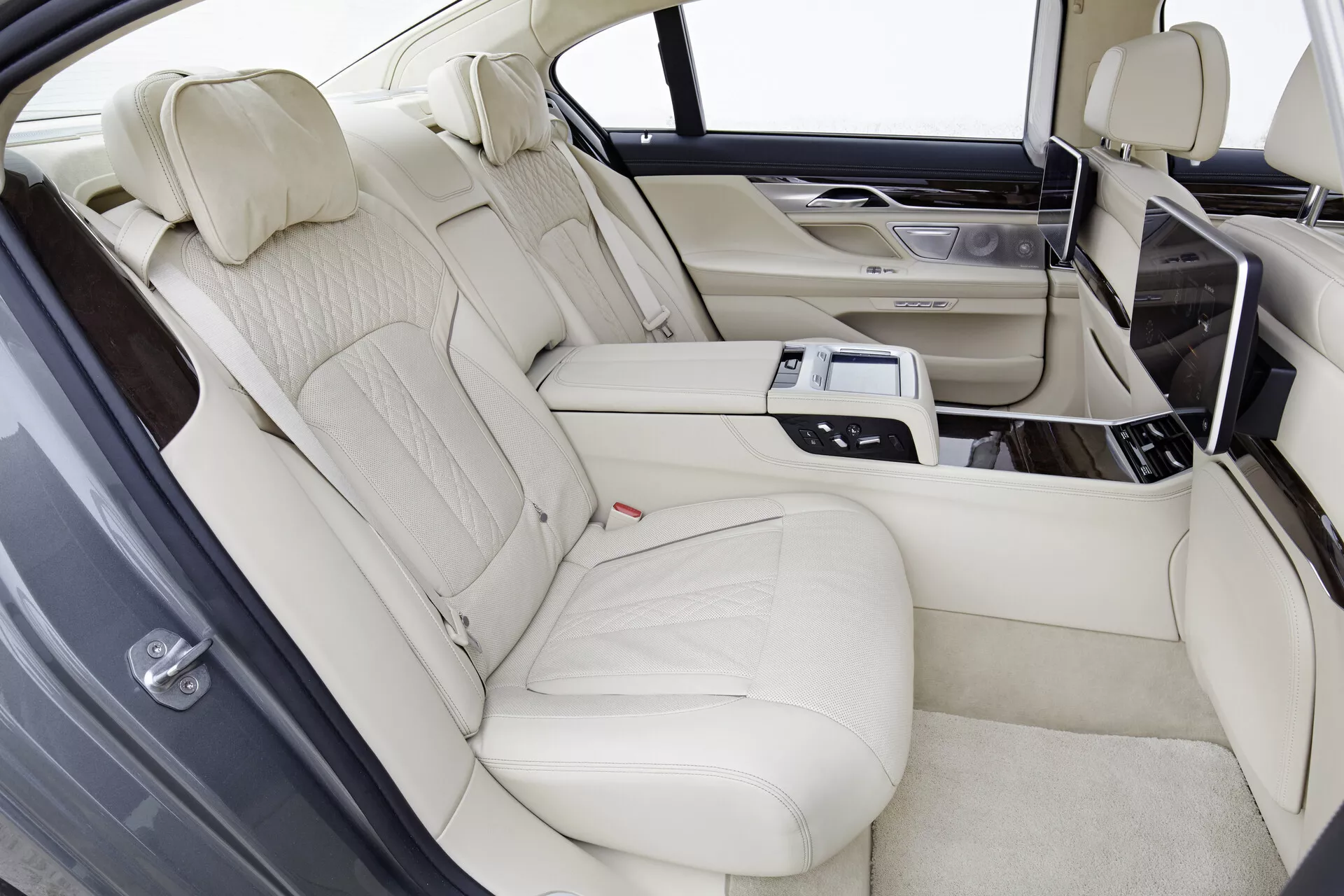 Wnętrze BMW serii 7 (G11-G12) model 2015, prezentujące luksusową tylnią przestrzeń siedzeń z jasnobeżową skórzaną tapicerką, profilowane tylne siedzenia dla dwóch pasażerów podzielone konsolą centralną. Konsola zawiera udogodnienia technologiczne, takie jak panel sterowania, klimatyzacja, ogrzewanie lub chłodzenie siedzeń, opcje multimedialne i uchwyty na kubki. Ekrany rozrywki tylnej są zintegrowane w tylnych częściach przednich siedzeń, z drewnianymi wykończeniami na konsoli i drzwiach, w jasnych warunkach oświetleniowych.