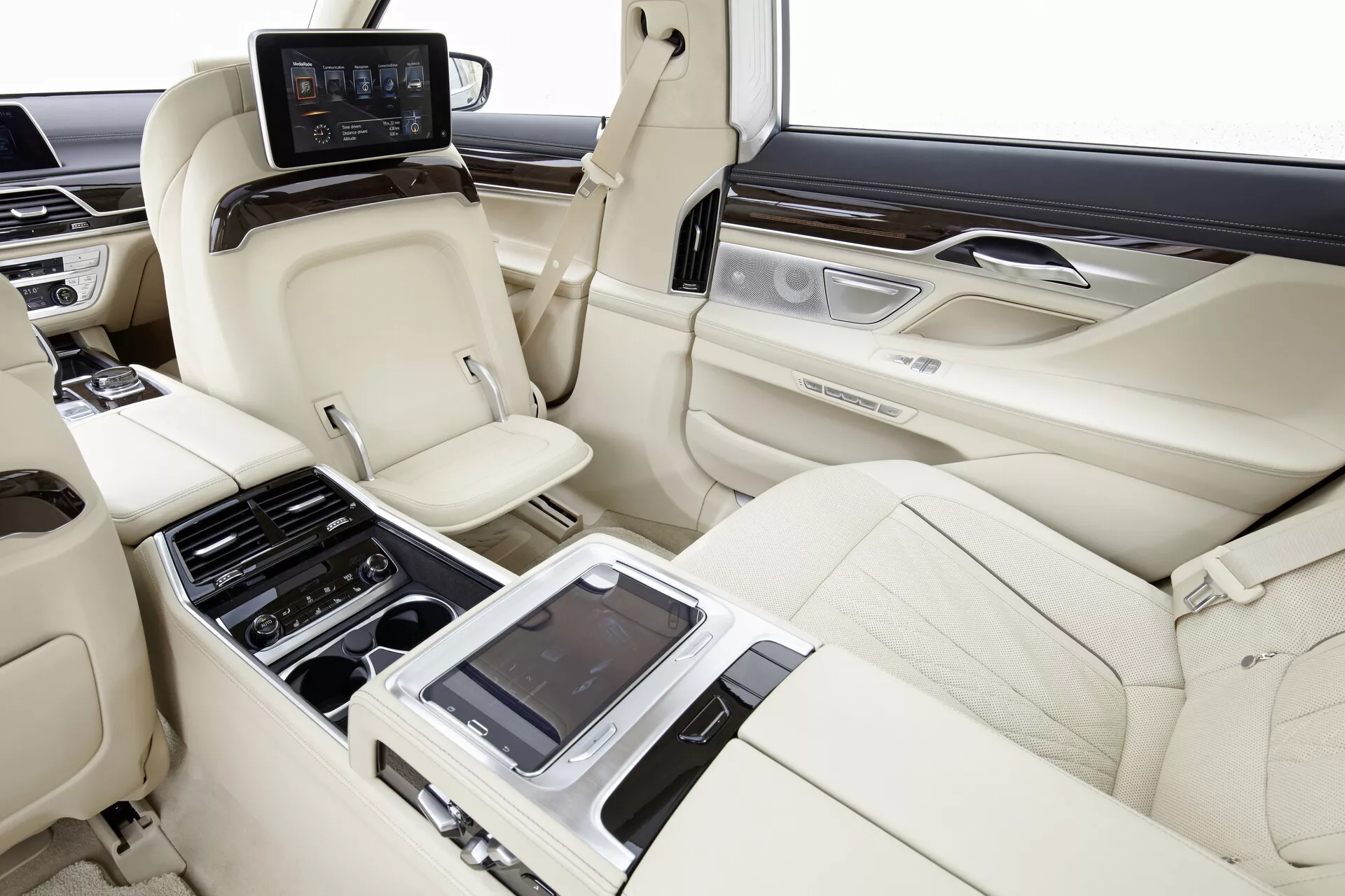 Obraz wnętrza kabiny tylnej modeli BMW 7er-reihe (G11-G12) z 2015 roku, ukazujący przestronną i luksusową powierzchnię siedzeń z prawej strony. Skórzane siedzenia w jasnym kolorze w układzie 2+1, z profilowanymi siedzeniami dla komfortu i wsparcia, zagłówki i indywidualne możliwości pochylania są widoczne. Centralna konsola z różnymi przyciskami sterującymi i prawdopodobnie interfejsem dotykowym do ustawień siedzeń, sterowania klimatyzacją oraz multimedia znajduje się w centrum. Panele drzwi zawierają elementy drewniane i szczotkowane metalowe akcenty z dodatkowymi przyciskami. Indywidualne monitory są zamontowane na tylnej części przednich siedzeń dla rozrywki, a panel kontrolny z ekranem znajduje się za centralną konsolą. Wnętrze pojazdu pokazuje zaawansowane ustawienia siedzeń, kontrolę klimatyzacji oraz systemy rozrywkowe w finiszach z wysokiej jakości skóry, drewna i metalu.