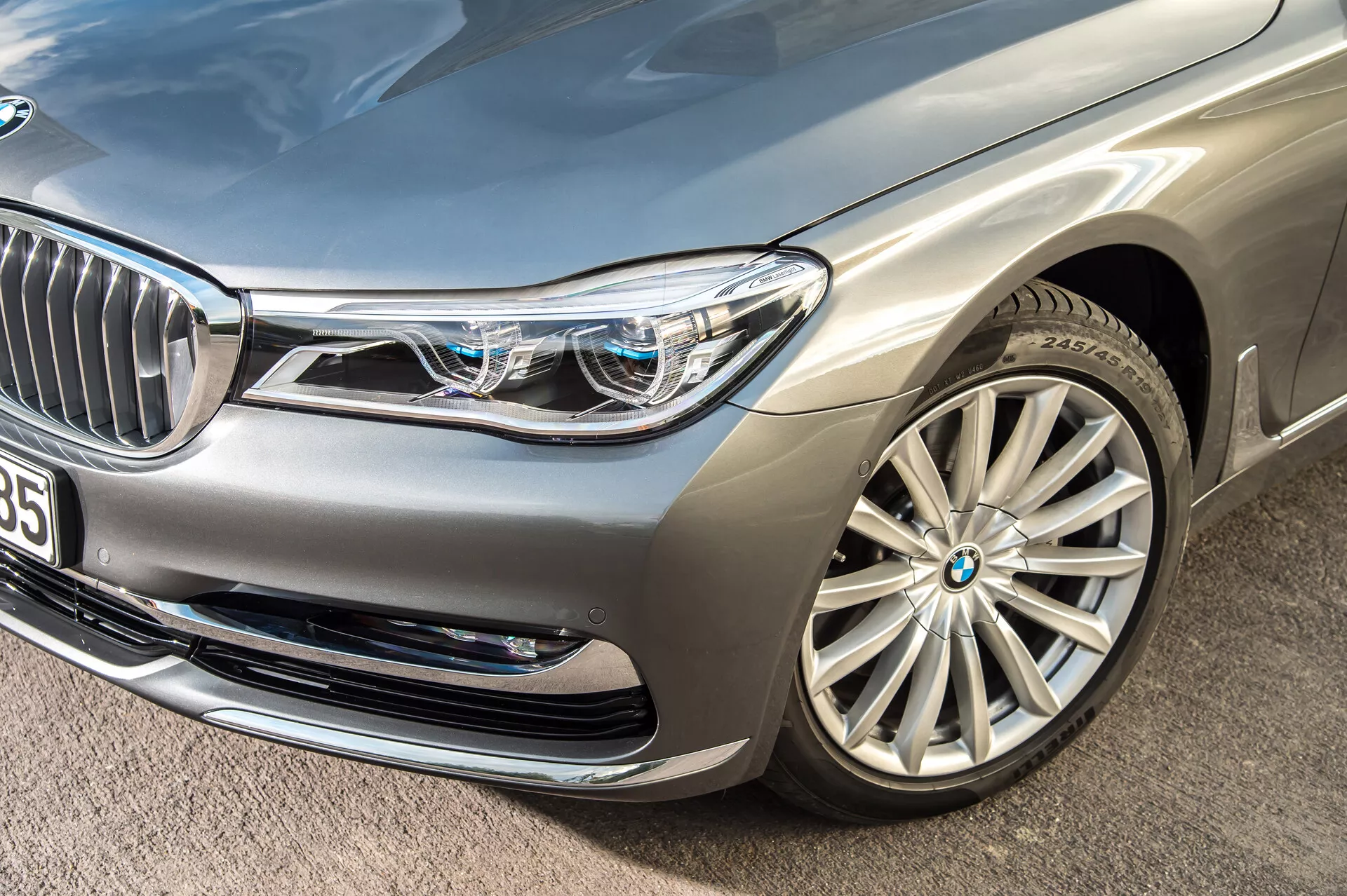 Zbliżenie pod kątem na srebrny luksusowy sedan BMW 7er-reihe 2015 (G11-G12), koncentrujący się na przedniej lewej części samochodu. Podkreślone są przedni zderzak, lewy reflektor, maska, błotnik i wieloszprychowe aluminiowe koło. Złożone reflektory LED z charakterystycznymi światłami do jazdy dziennej i małymi niebieskimi kwadratami wskazującymi zaawansowane systemy oświetleniowe. Częściowo widoczna przednia krata z chromowanymi pionowymi listewkami i pobliską kamerą lub czujnikiem sugerują technologię wspomagania kierowcy. Wloty powietrza w przednim zderzaku i metaliczne wykończenie lakieru dodatkowo podkreślają luksusowy i aerodynamiczny design samochodu.