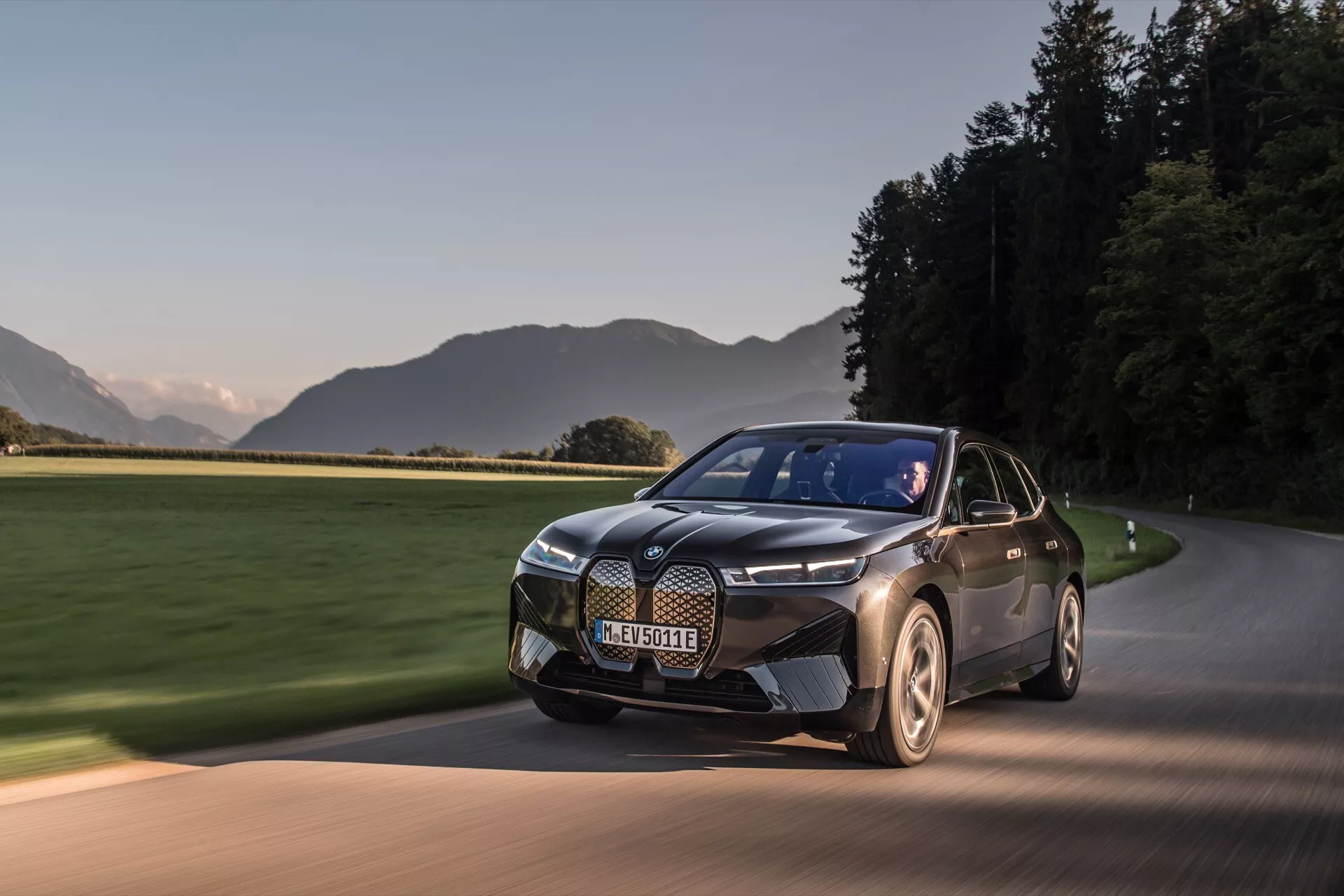Nowoczesny elektryczny SUV BMW iX 2021 (i20) w ruchu na utwardzonej drodze o świcie lub zmierzchu, prezentujący dwukolorowe malowanie, elegancki design z wyraźną kratą, charakterystyczne reflektory i aerodynamiczne koła na tle malowniczego krajobrazu.