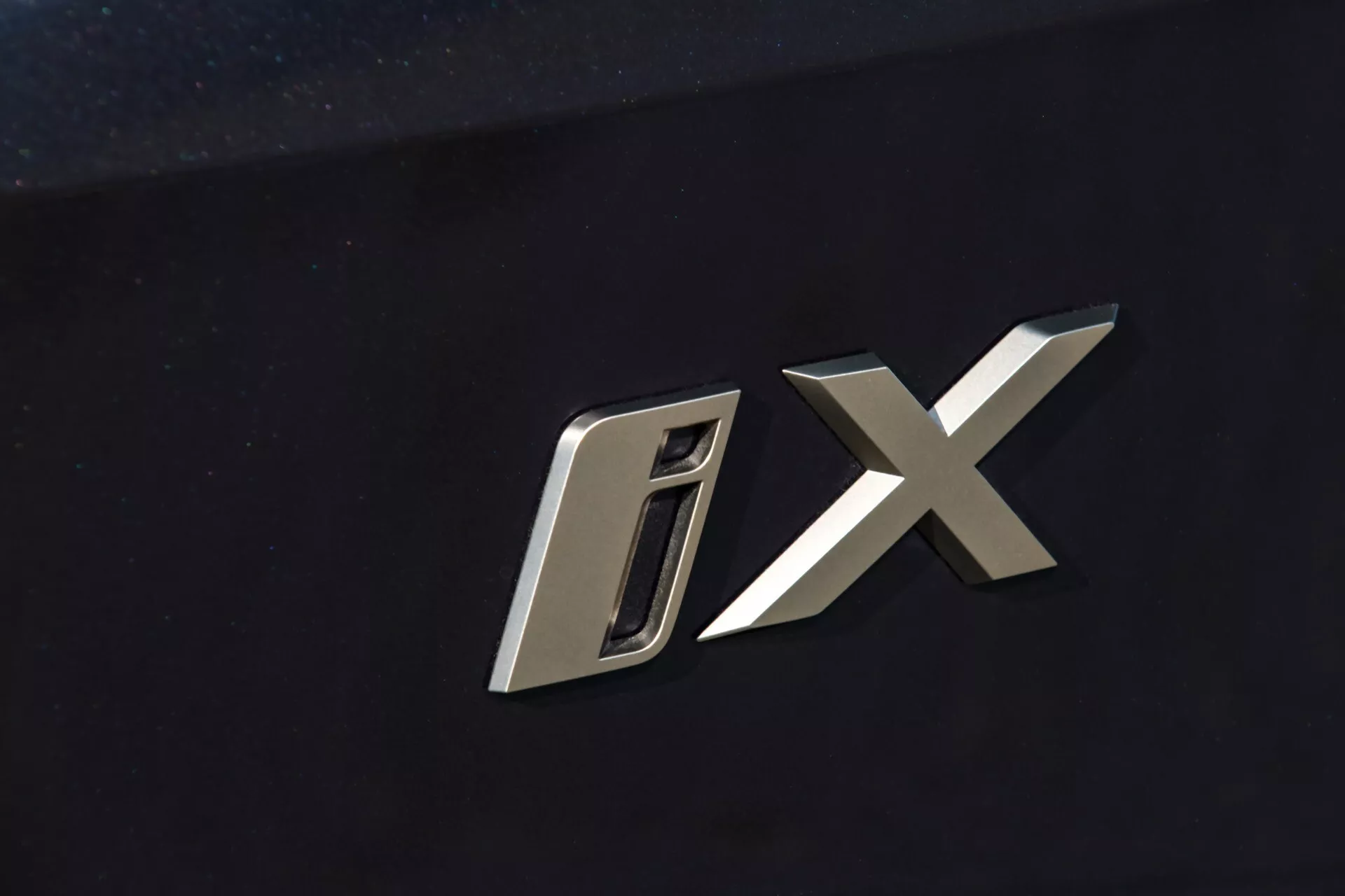 Zbliżenie na emblemat modelu BMW iX 2021 z stylizowanymi metalicznymi literami 'D' i 'X' na ciemnym tle karoserii