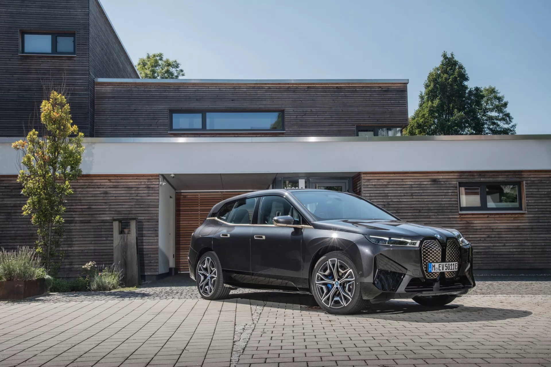 BMW iX 2021 (i20) zaparkowane na podjeździe, pokazuje perspektywę przednio-boczną z budynkiem mieszkalnym w tle, z agresywnym przednim grillem, smukłymi reflektorami LED i dużymi kołami ze stopu o wielu ramionach.