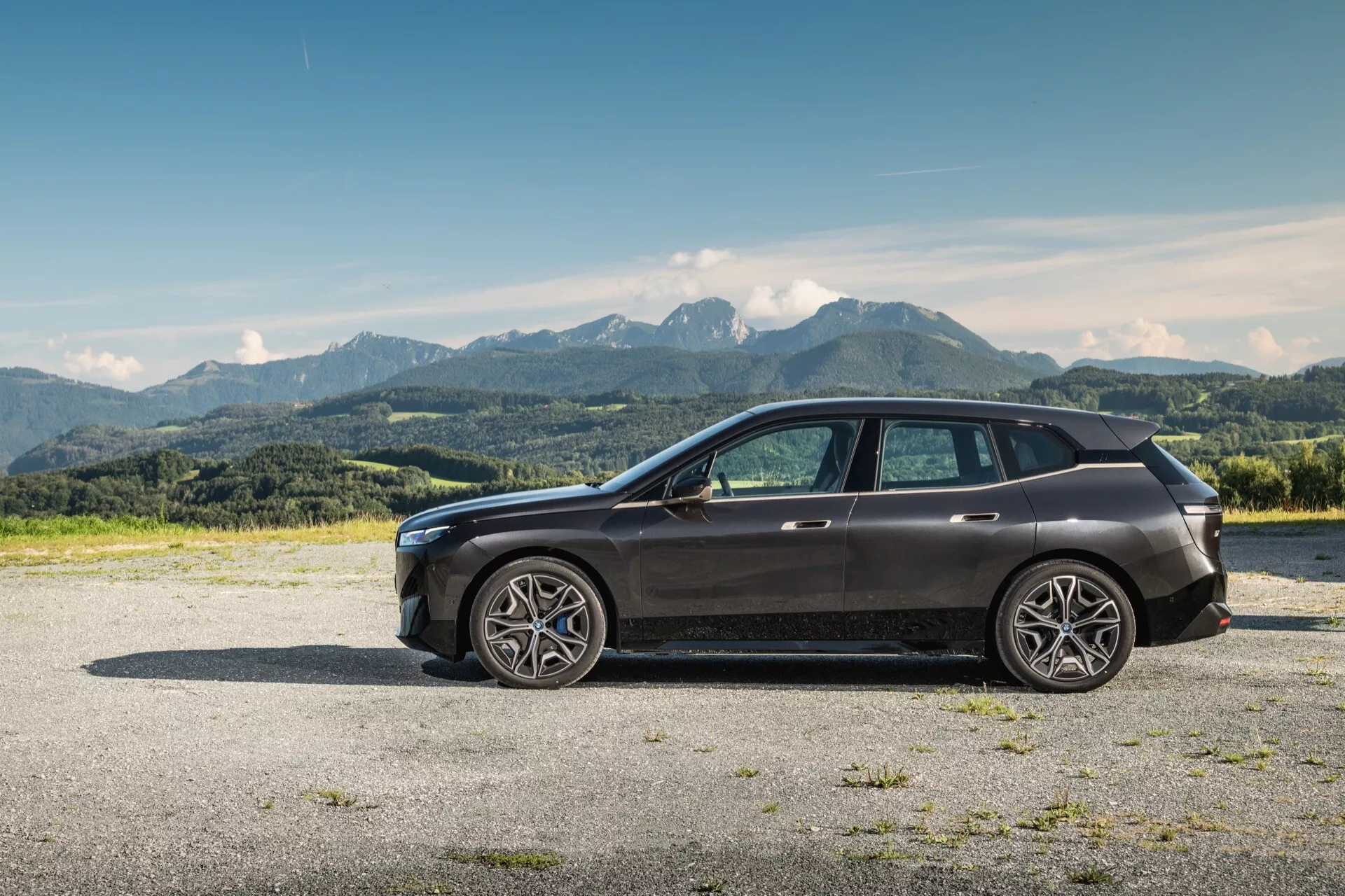 Boczny profil nowoczesnego SUV BMW iX (i20) z 2021 roku na żwirze z górskim tłem pod czystym błękitnym niebem, z dużymi kołami ze stopu o wielu szprychach, eleganckim designem z wysoką linią pasa i opadającym dachem, szkłem zapewniającym prywatność, wbudowanymi w lusterka w kolorze karoserii kierunkowskazami, widoczną przednią kratą i zespołem reflektorów LED, chromowanymi wykończeniami i panoramicznym dachem.