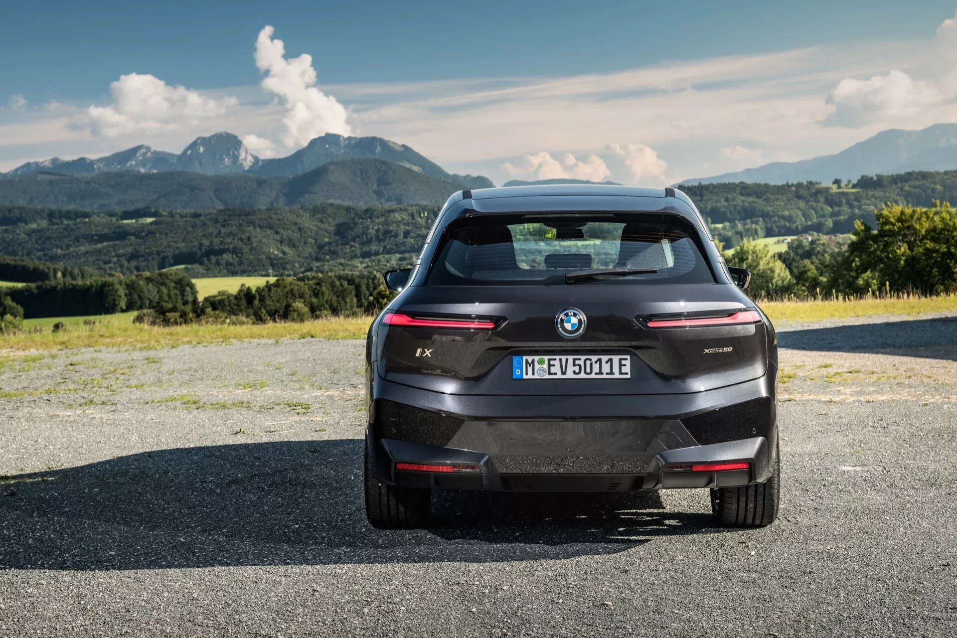 Tylny widok BMW iX 2021 SUV elektrycznego na asfaltowej powierzchni z górami i częściowo pochmurnym niebem w tle, prezentujący nowoczesny design, eleganckie linie, logo BMW i oznaczenie modelu, zintegrowane czujniki parkowania z tyłu, wysoko zamontowane światło stop, kamerę cofania, smukłe poziome tylne światła i aerodynamiczne tylne okno bez tradycyjnego wydechu, wskazujące na elektryczny charakter pojazdu i rejestrację w Monachium.