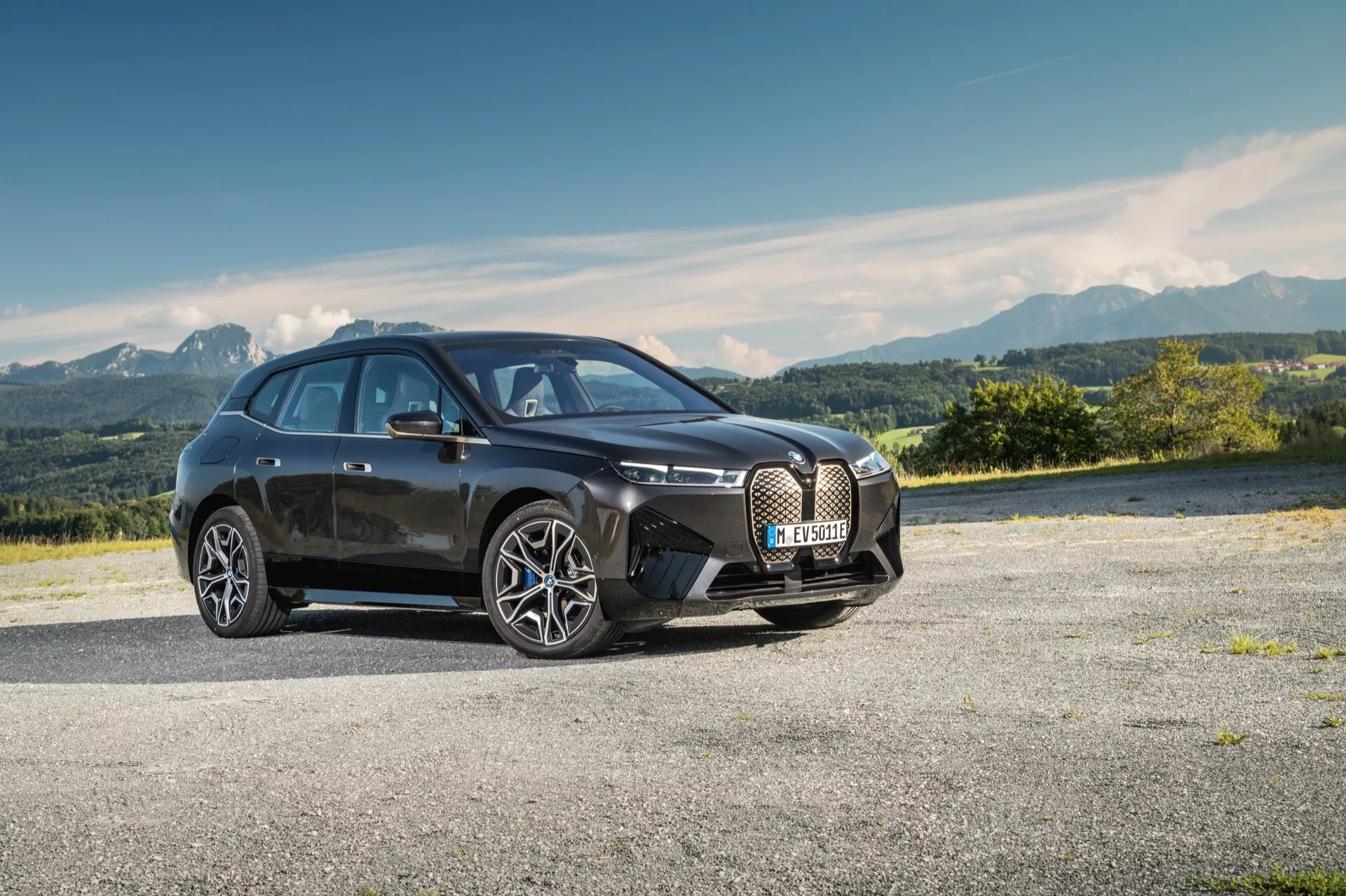 Nowoczesny hatchback czterodrzwiowy BMW iX (i20) z 2021 roku zaparkowany na wybrukowanym obszarze z górskim krajobrazem w tle, prezentujący dużą atrapę chłodnicy w kształcie nerki, eleganckie linie, reflektory LED i aerodynamiczny design z dużymi kołami ze stopu