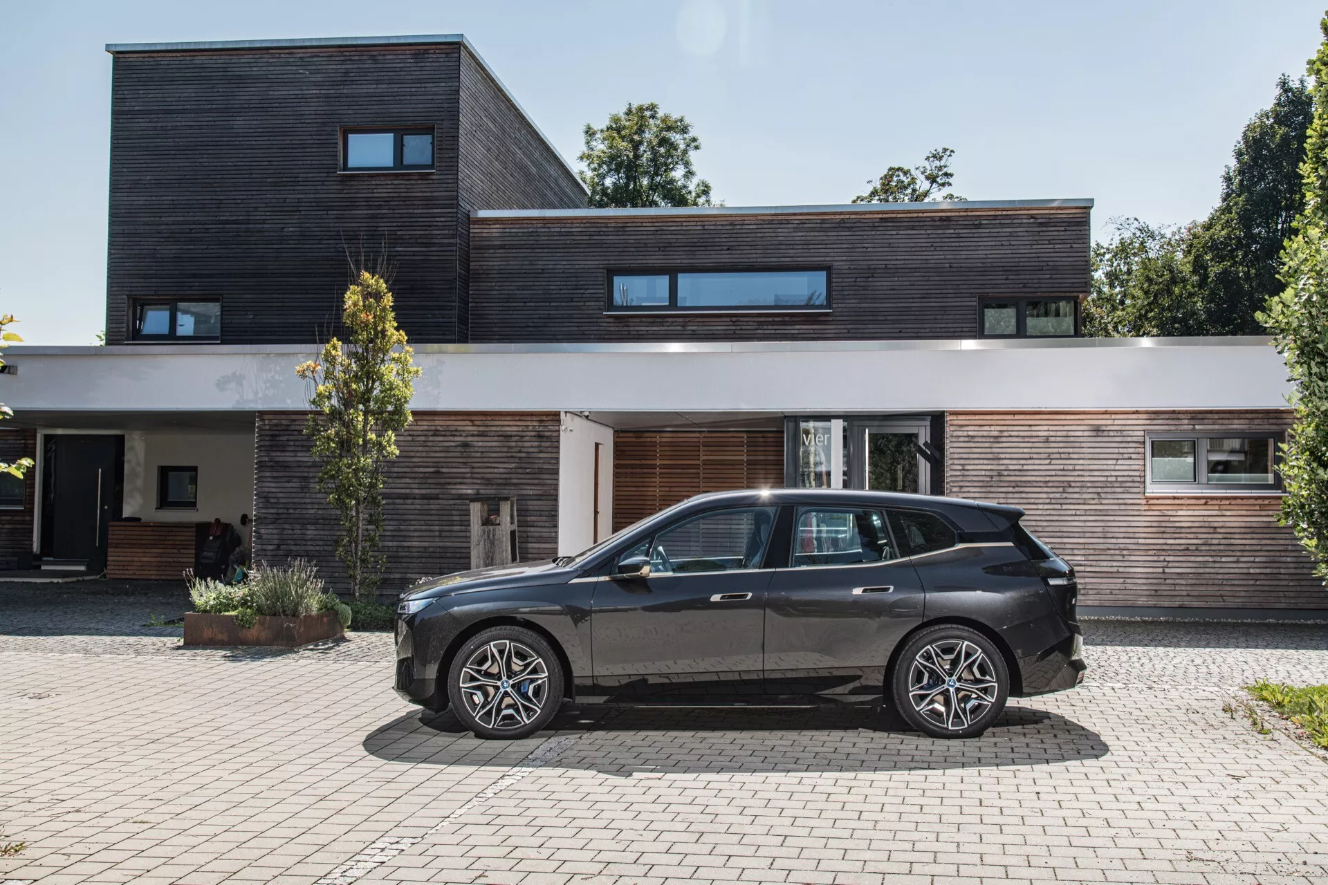 BMW iX (i20) z 2021 roku zaparkowane przed nowoczesnym domem z okładziną drewnianą i ciemnymi materiałami, prezentujące boczny profil samochodu, aerodynamiczny design, felgi aluminiowe i wysoki prześwit