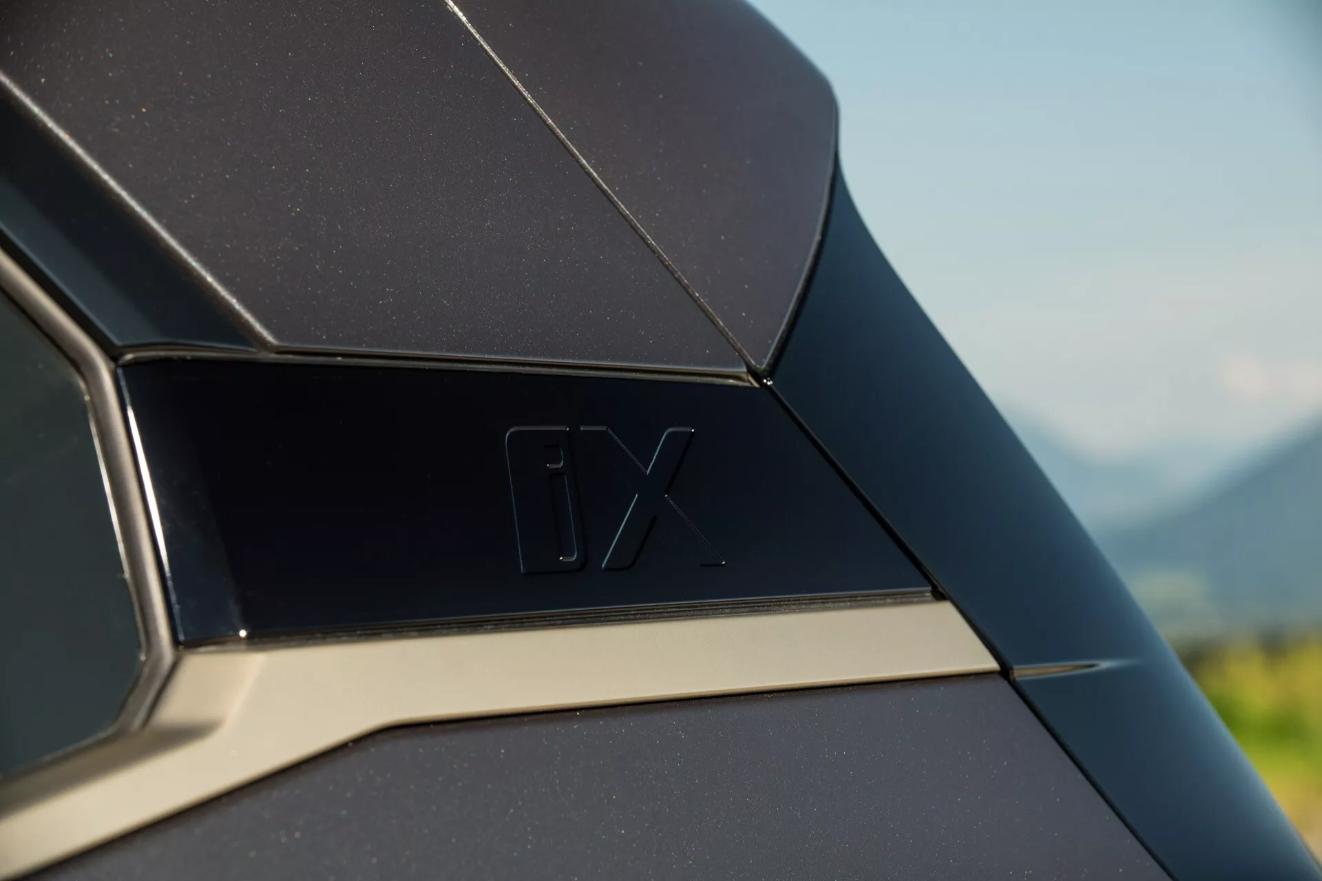 Zbliżenie na szczegół zewnętrzny BMW iX 2021, przedstawiające tylny panel boczny lub obszar słupka C z emblematem 'iX' i kontrastującą chromowaną listwą, na rozmytym tle naturalnym