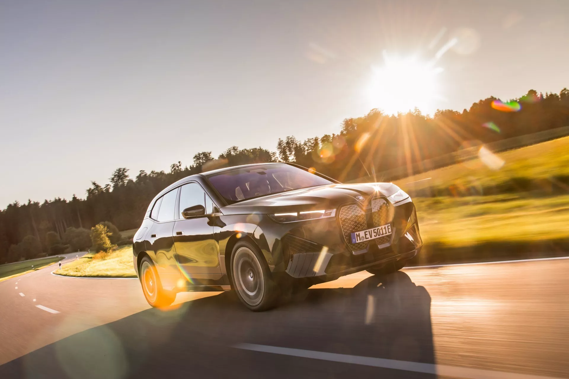 Dynamiczny widok z przodu pod kątem trzech czwartych BMW iX 2021 w ruchu, podkreślający sportową postawę, eleganckie reflektory LED i charakterystyczną dużą kratkę, uchwycony z niskiego kąta, aby podkreślić nowoczesne i agresywne cechy projektu samochodu na tle naturalnego wiejskiego krajobrazu.