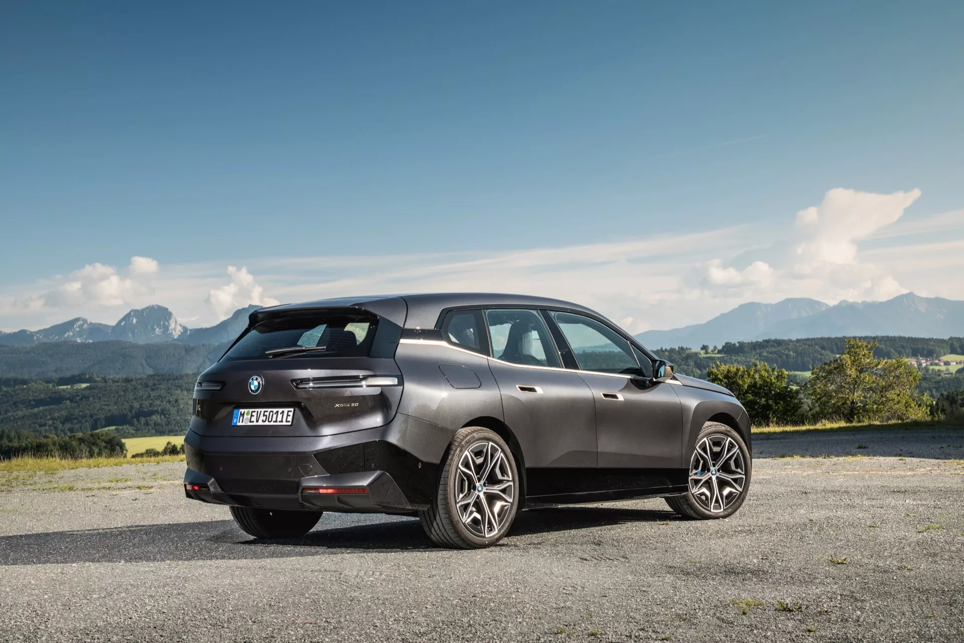 Widok z tyłu i boku na BMW iX 2021 w ciemnoszarym kolorze, zaparkowany na podwyższonej nawierzchni z górskim krajobrazem w tle, odzwierciedlający nowoczesny design luksusowego SUV-a