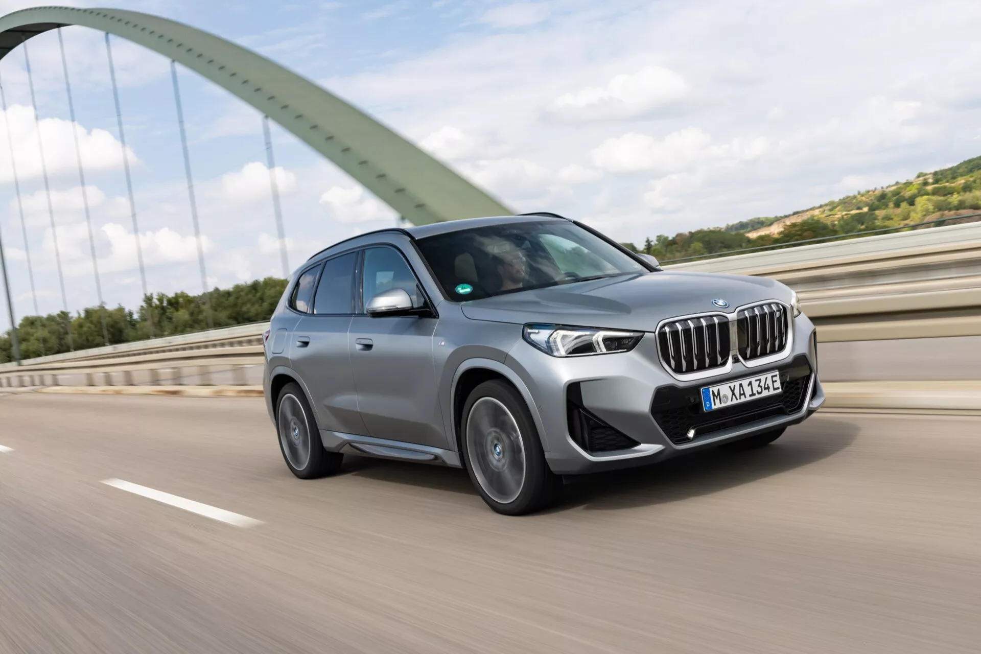 Nowoczesne BMW iX1 2022 (U11) SUV w ruchu uchwycony z widoku przedniego trzech czwartych na wyniesionej drodze lub moście. Zdjęcie podkreśla charakterystyczną osłonę chłodnicy z aktywnym systemem żaluzyjnym, wąskie i eleganckie reflektory LED, zintegrowane światła przeciwmgielne i wloty powietrza na przednim zderzaku, możliwe że wyposażone w czujniki do systemów wspomagania kierowcy. Niebieskie akcenty wokół osłony chłodnicy i odznaka elektryfikacji wskazują, że jest to model zelektryfikowany. Obręcze ze stopów metali lekkich z wieloramiennym wzorem i widocznym bieżnikiem opon całorocznych sugerują sportową wersję. Tło z zielonym łukowym mostem dodaje kontrastowy kolor, podkreślając zaawansowane technologie i dynamiczne osiągi tego nowego modelu.