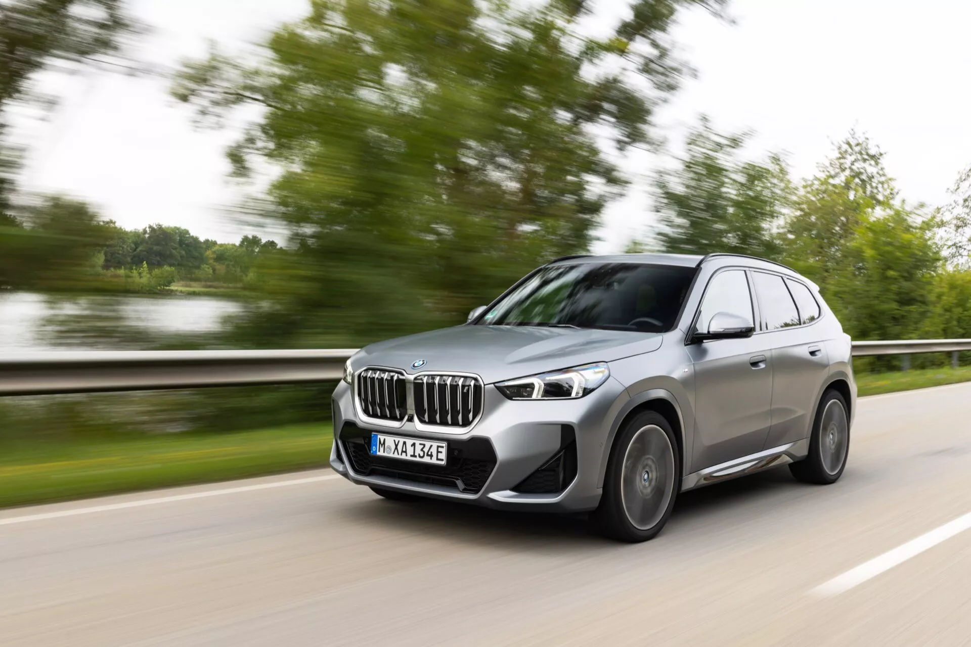 Obraz BMW iX1 2022 (U11), nowoczesnego SUV-a średniej wielkości w ruchu uchwycony z przedniego trzech czwartych kąta, ukazujący przód, stronę kierowcy i przednie koło z charakterystycznym grillem nerkowym, eleganckimi reflektorami LED i dużymi wlotami powietrza. Aluminiowe felgi, szary kolor, dynamicznie zaprojektowane lusterka boczne i rozmyte tło podkreślają prędkość i ruch SUV-a, co wskazuje na model hybrydowy lub zelektryfikowany.
