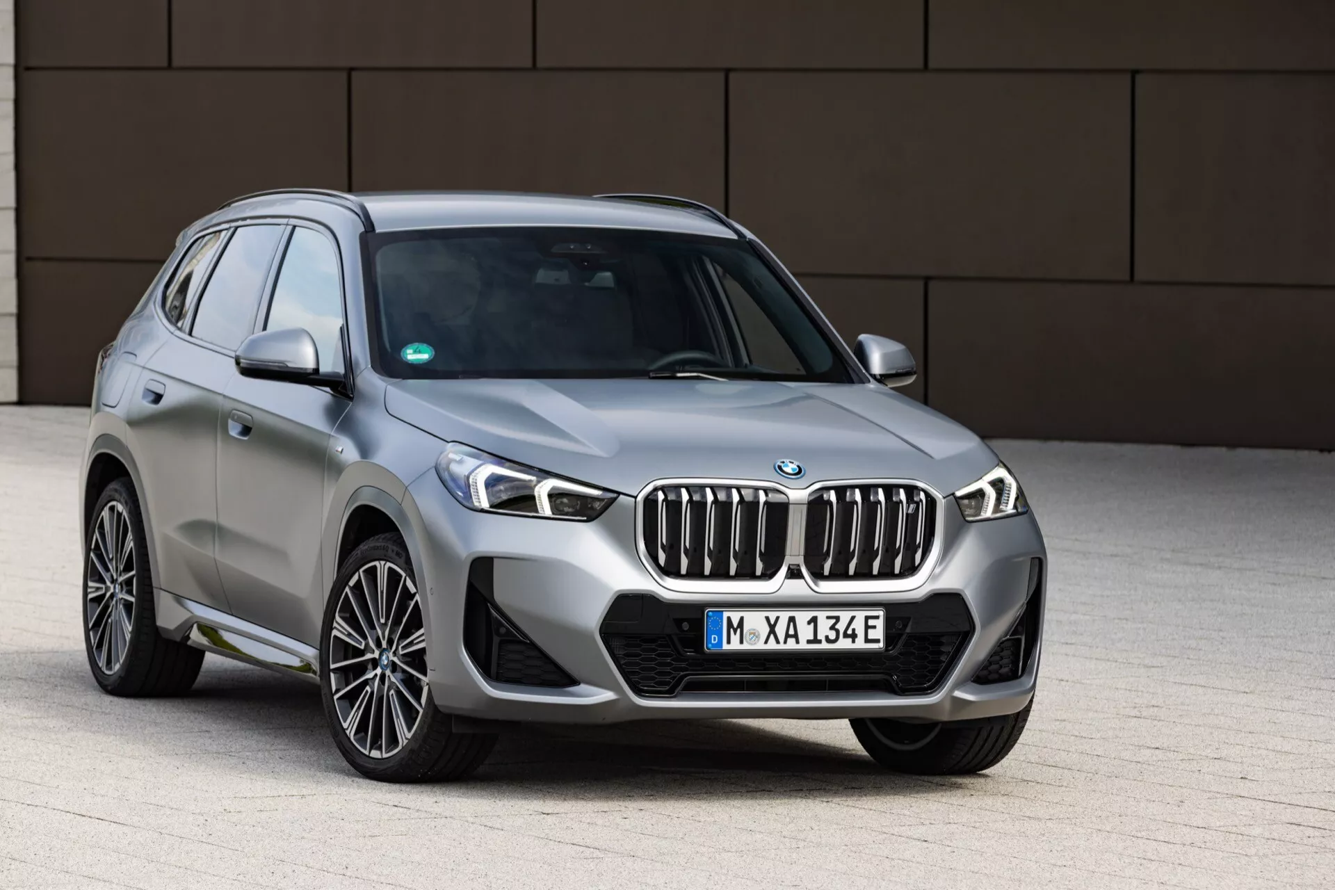 Trzy czwarte przedniego widoku nowoczesnego SUV-a BMW iX1 2022 (U11), pokazując charakterystyczną nerkową kratkę, eleganckie reflektory LED z wydłużonymi heksagonalnymi światłami do jazdy dziennej, przedni zderzak, maskę i lewe koła oraz błotniki. SUV sugeruje model hybrydowy lub elektryczny, oznaczony niebieskimi akcentami wokół emblematu kratki, 'E' na tablicy rejestracyjnej i klapą portu ładowania na przednim błotniku. Uchwycony na poziomie oczu, pojazd stoi na brukowanej powierzchni z minimalistycznym tłem, oświetlenie jest miękkie i równomierne, podkreślając metaliczne wykończenie pojazdu i wypolerowane opony.