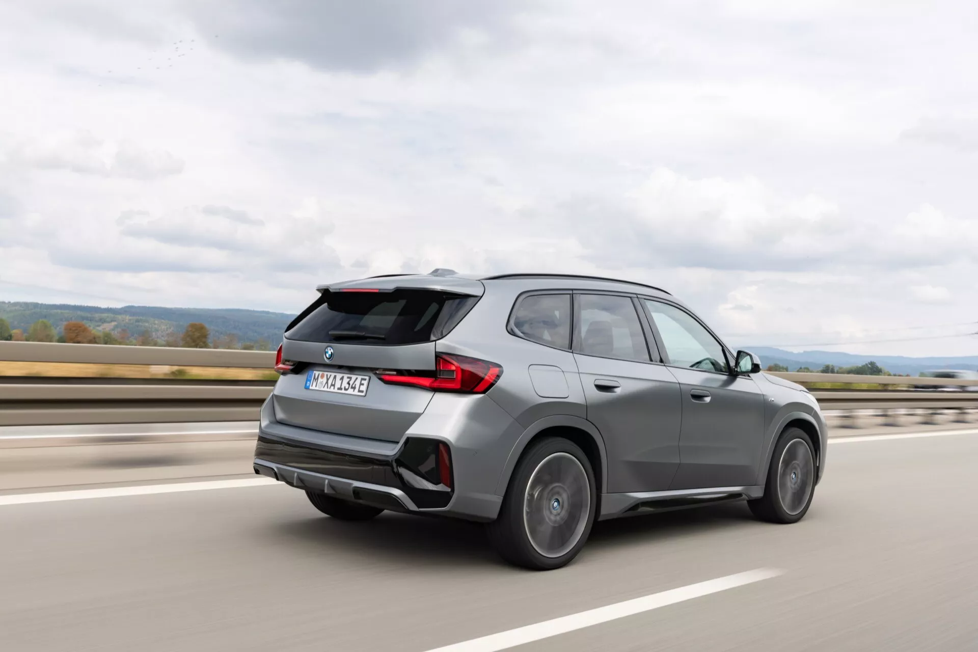 Obraz przedstawia SUV BMW iX1 2022 (U11) w ruchu na autostradzie, uchwycony z tyłu z trzy czwarte widoku. Nowoczesny design pojazdu charakteryzuje się rzeźbionymi liniami nadwozia, zintegrowanym tylnym spojlerem, nowoczesnymi tylnymi światłami LED, podwójnymi końcówkami wydechu, charakterystycznym wzorem kół, przyciemnianymi szybami tylnymi i dyfuzorem tylnym. Rozmycie wokół kół i tła wskazuje, że SUV porusza się z dużą prędkością.