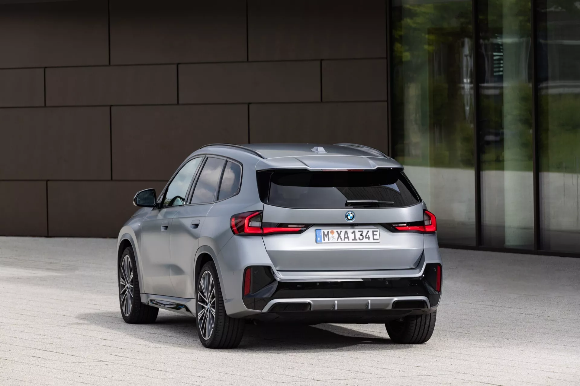 Perspektywa tylna trzy czwarte nowoczesnego kompaktowego SUV-a, uwydatniająca tył i bok. Cechy obejmują dwuczęściowe tylne światła LED rozciągające się na klapę bagażnika, centralnie umieszczony emblemat producenta, subtelny tylny spojler i szary dyfuzor z poziomymi elementami na tylnym zderzaku. Boczne kontury kierowcy pokazują aerodynamiczne kontury i wpuszczane klamki. Koła ze stopów mają wieloramienny wzór, który sugeruje nacisk na estetykę i aerodynamikę. Brak widocznych wylotów wydechowych wskazuje na napęd elektryczny lub hybrydowy.