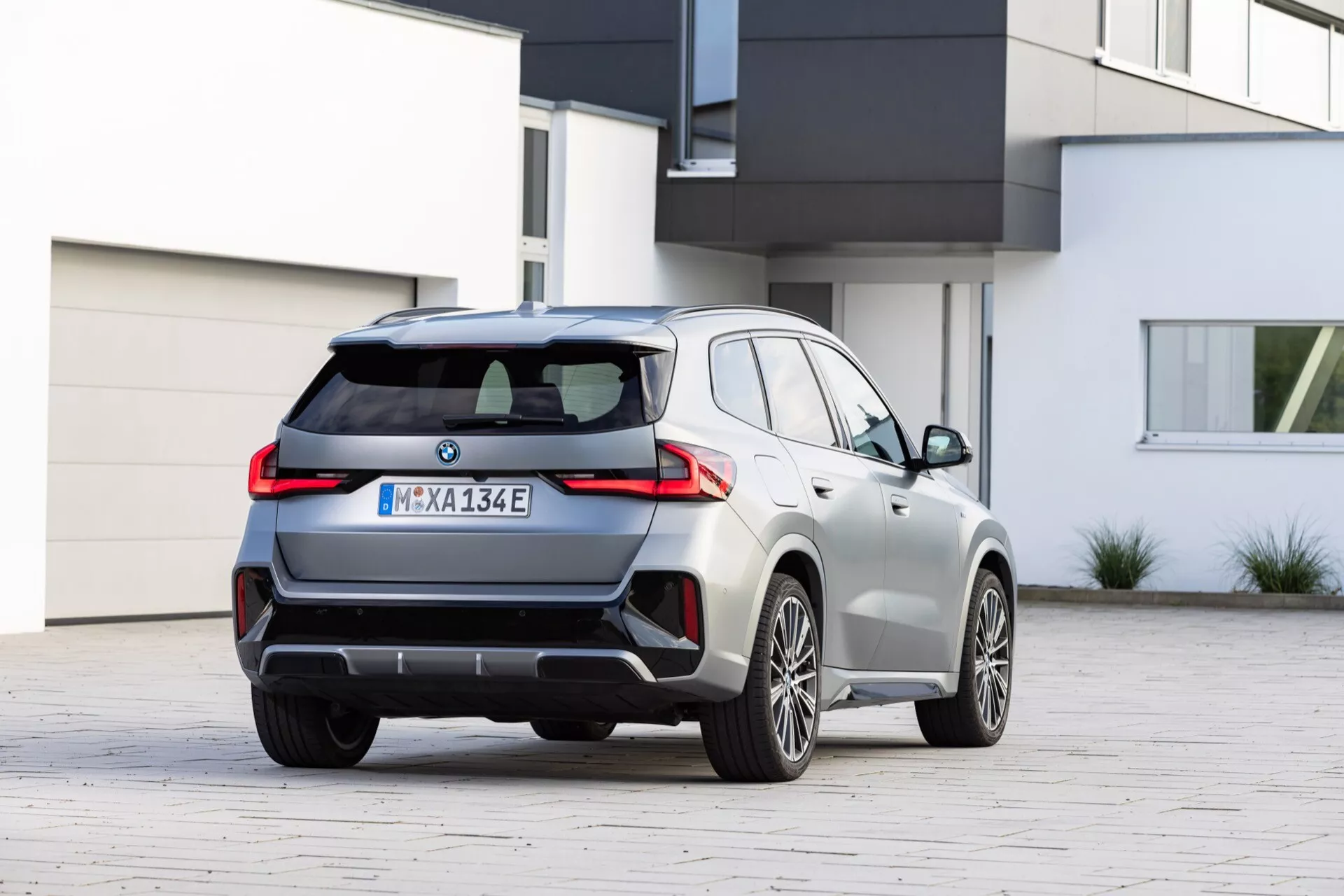 Średniej wielkości luksusowy crossover SUV w widoku tylnym trzy czwarte, pokazującym tył i prawą stronę. Widoczne są bagażnik, tylny zderzak, tylne światła o wyglądzie LED, podwójny wydech, linia barku, klamki i wieloramienne felgi aluminiowe oraz metaliczne srebrzyste wykończenie. Rozmyte logo sugeruje model elektryczny lub hybrydowy.