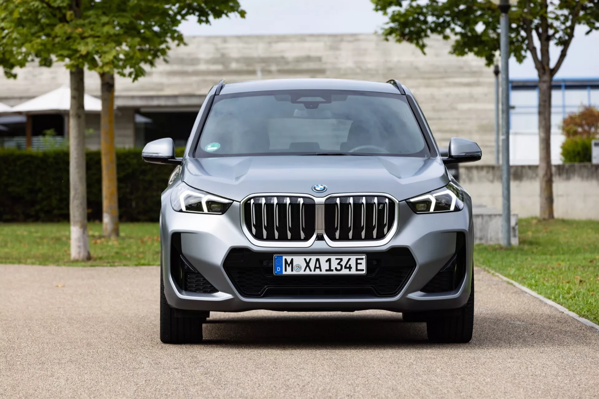 Widok z przodu BMW iX1 2022 (U11), ukazujący charakterystyczną nerkowatą kratkę chłodnicy otoczoną kanciastymi reflektorami LED z DRL, wyrazisty dolny zderzak z światłami przeciwmgielnymi i wlotami powietrza, widocznymi czujnikami parkowania i centralnym znaczkiem wskazującym na elektryczną/hybrydową wersję.
