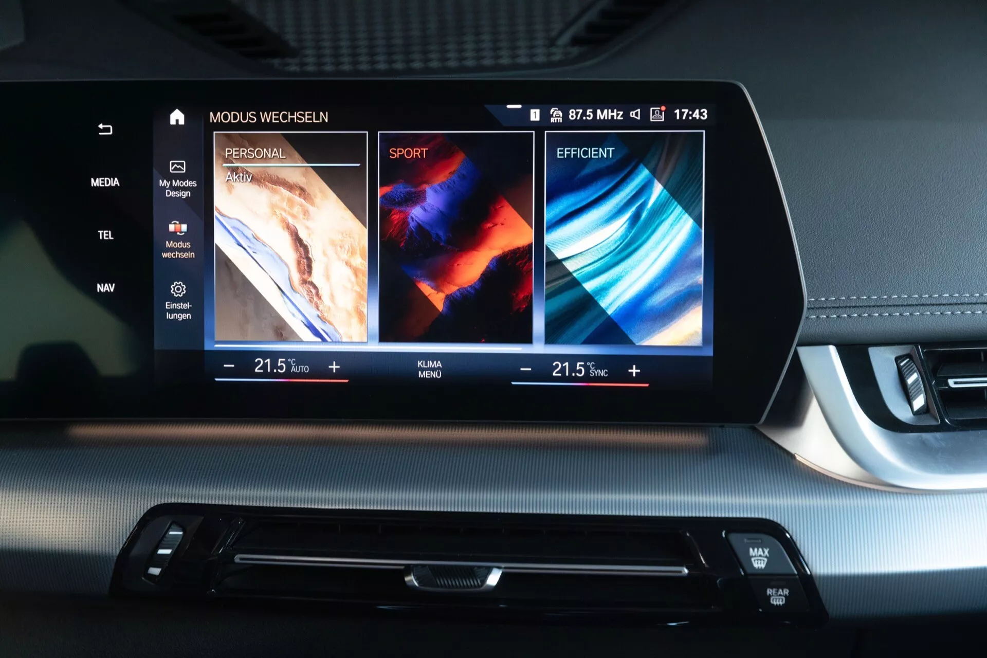 Profesjonalne zdjęcie wnętrza BMW iX1 2022 (U11) z perspektywy kierowcy lub pasażera przedniego, przedstawiające szerokoekranowy system multimedialny. Ekran zawiera sekcje Media, Telefon i Nawigacja oraz konfigurowalne tryby jazdy - Personal, Sport i Efficient. Poniżej ekranu deska rozdzielcza zawiera fizyczne elementy sterujące dla dwustrefowej klimatyzacji ustawionej na 21,5°C oraz szczelinę na odtwarzacz CD, co świadczy o nowoczesnym, technologicznym i luksusowym designie pojazdu.