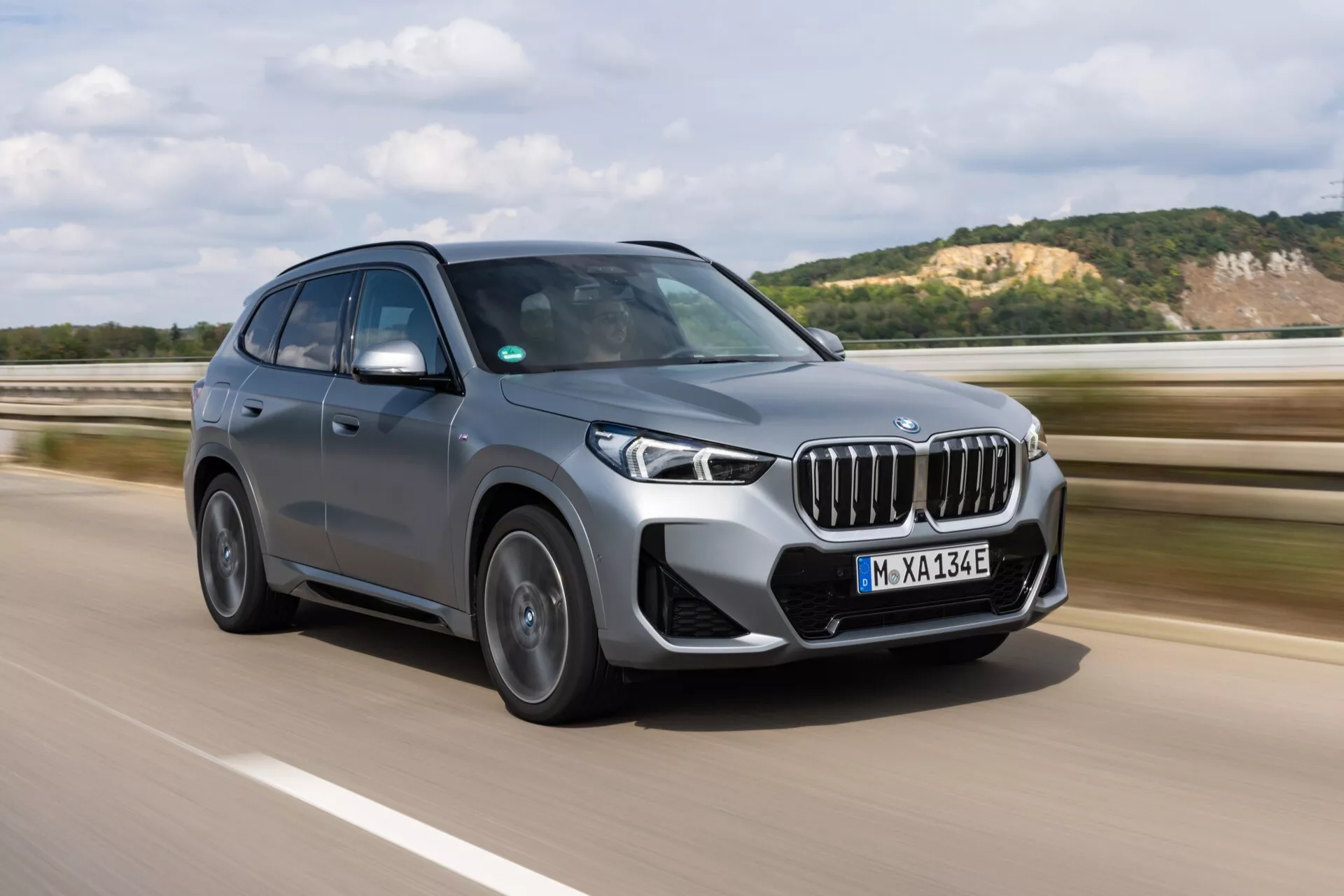 BMW iX1 2022 (U11) jest widoczny w ruchu na autostradzie, uchwycony z przedniego trójwymiarowego kąta. Obraz podkreśla przednią kratkę pojazdu, ostre reflektory LED i wyraźne elementy wzornictwa po stronie kierowcy. Dzienny strzał z naturalnym oświetleniem podkreśla metaliczne wykończenie lakieru SUV-a, aerodynamiczne linie i wieloramienne felgi aluminiowe. Przyciemniane szyby i lusterka boczne w kolorze nadwozia z zintegrowanymi kamerami lub wskaźnikami sugerują zaawansowane technologie wspomagania kierowcy. Elektryczny charakter pojazdu jest oznaczony znakiem 'Electric Test Vehicle' oraz plakietką modelu. Tło jest lekko rozmyte, aby podkreślić dynamiczne osiągi pojazdu.