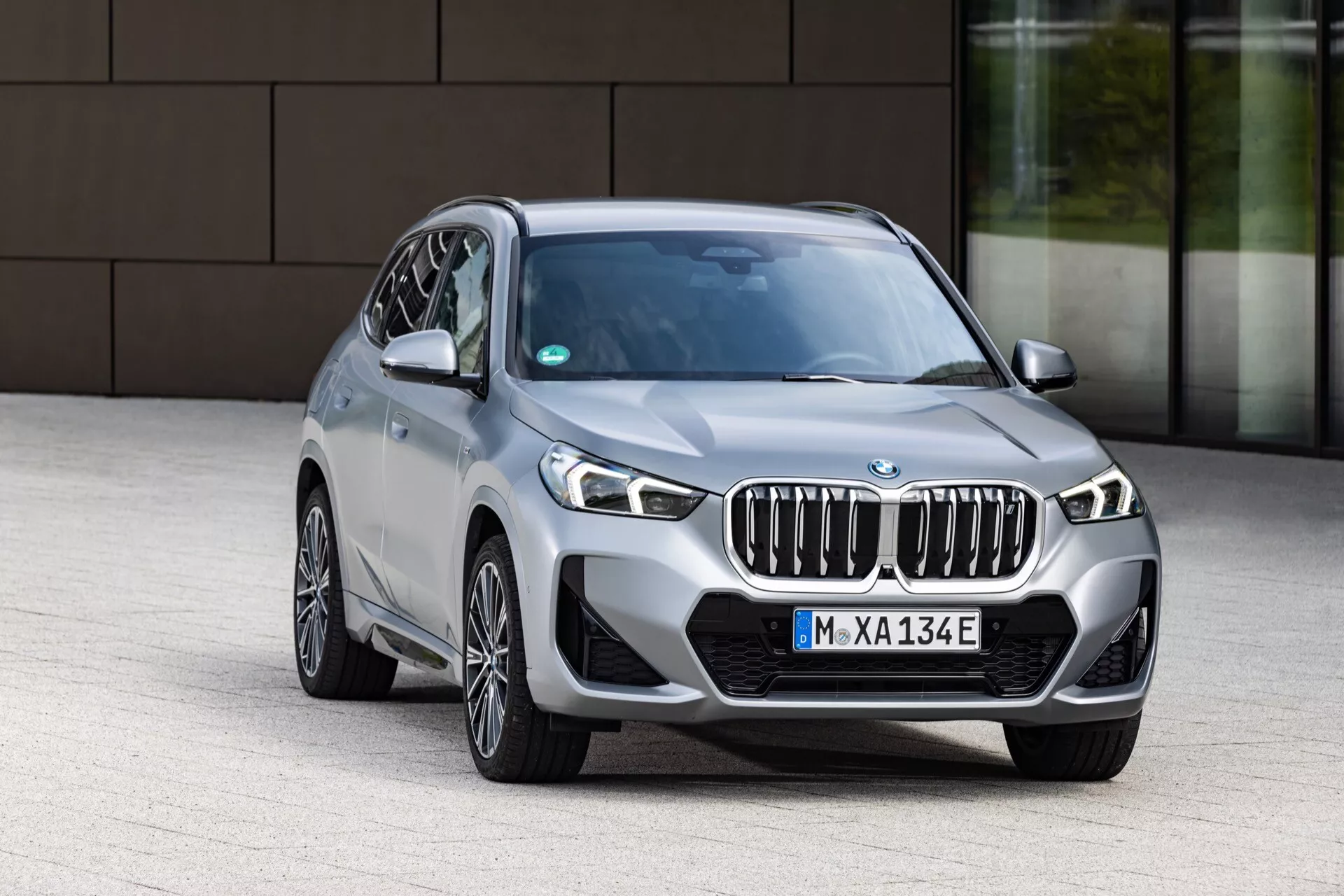 Widok z przodu pod kątem trzech czwartych luksusowego SUV-a BMW iX1 2022 (U11), podkreślający elementy designu, takie jak wyróżniająca się przednia osłona z pionowymi żaluzjami, skomplikowane reflektory LED, dolny zderzak z światłami przeciwmgielnymi lub wlotami powietrza oraz wieloramienne felgi aluminiowe w metalicznym wykończeniu. Samochód jest lekko przesunięty w lewo w sterowanym otoczeniu w pobliżu budynku korporacyjnego.