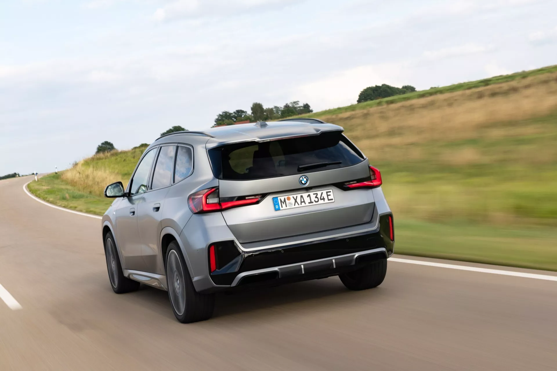 Dynamiczny widok z tyłu SUV-a BMW iX1 (U11) 2022 w ruchu, uchwycony z niskiego kąta, nieco przesunięty w prawo. Obraz podkreśla tylny design obejmujący tylne światła, końcówki wydechowe, zderzak i odznaczenia, z rozmyciem ruchu na kołach i tle. Zawiera tylny dyfuzor, czujniki lub kamery sugerujące system ADAS. Pokazuje sportowy, ale solidny design SUV-a z pochylonym dachem, relingami dachowymi i szarym lub srebrnym kolorem. Emblemat 'E' i oznaczenie 'Electric Test Vehicle' wskazują na elektryczny lub hybrydowy model.