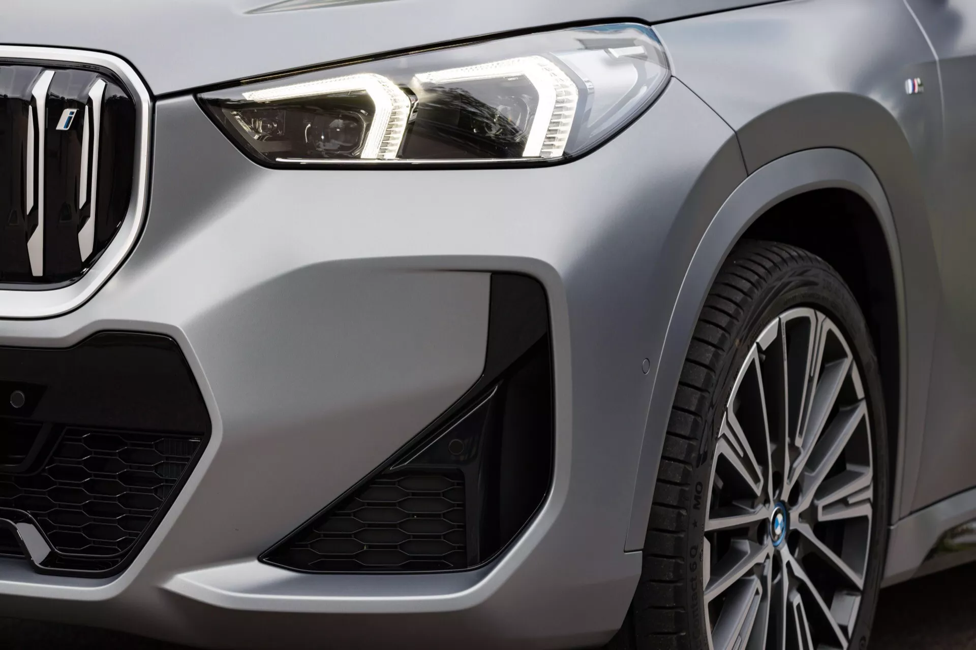 Zbliżenie pod kątem ukośnym na BMW iX1 2022 (U11), ukazujące nowoczesny reflektor LED, częściowo widoczną grilla z charakterystycznym emblematem, sportowy przedni zderzak z dużymi wlotami powietrza i fragment wieloszprychowego koła aluminiowego z przodu. Obraz podkreśla odważne linie i krzywizny lewej przedniej strony pojazdu, podkreślając jego luksusowe i sportowe elementy designu.