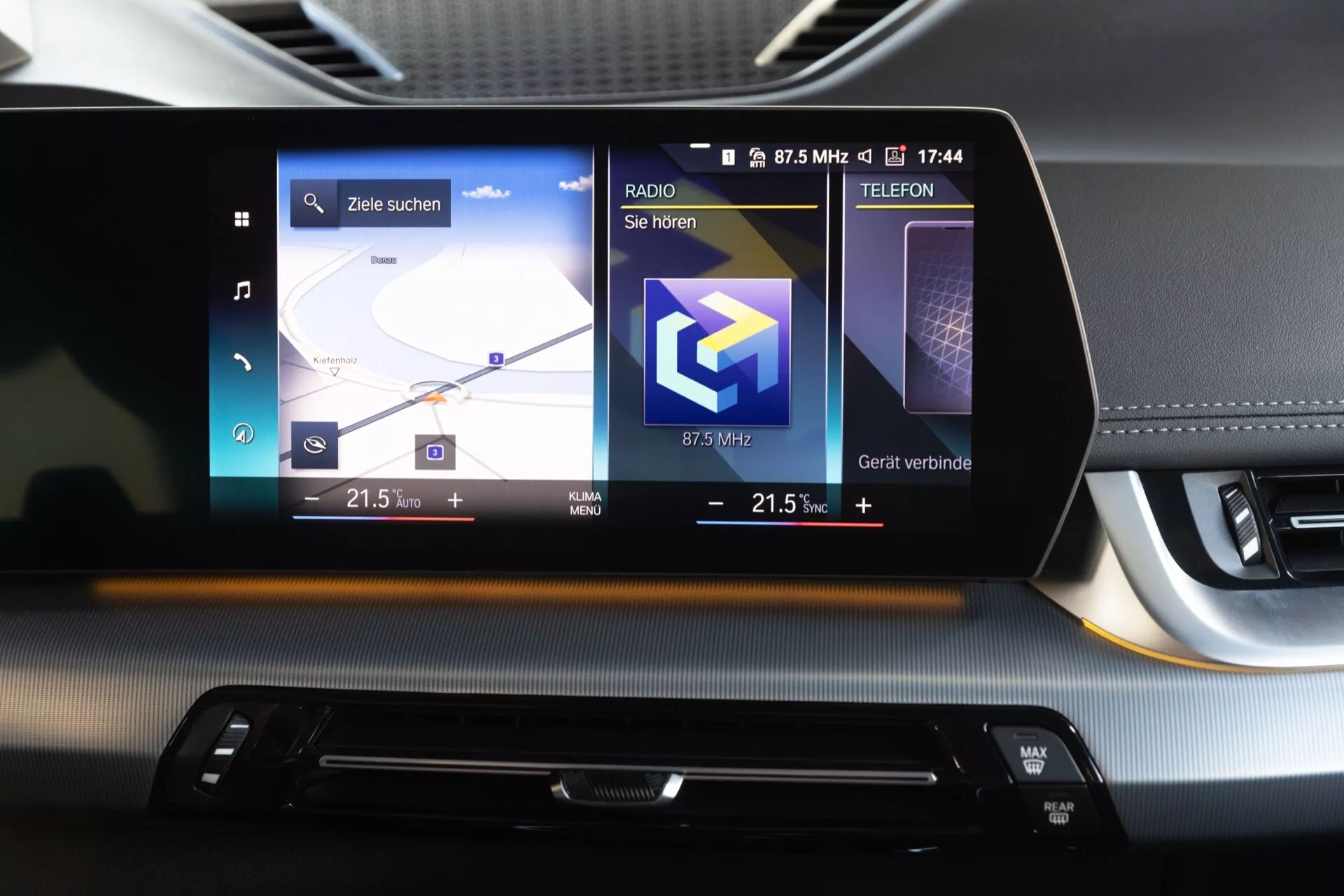 Zbliżenie na system infotainment i deskę rozdzielczą w BMW iX1 2022 (U11). Dotykowy ekran w orientacji poziomej pokazuje mapę nawigacji po lewej, stację radiową pośrodku i opcje łączności po prawej. Pod ekranem znajduje się wąski nawiew i elegancki panel sterowania dotykowego z dwustrefową klimatyzacją. Minimalistyczny design łączy teksturowane i gładkie materiały deski rozdzielczej, odzwierciedlając wysoką jakość wykonania samochodu.