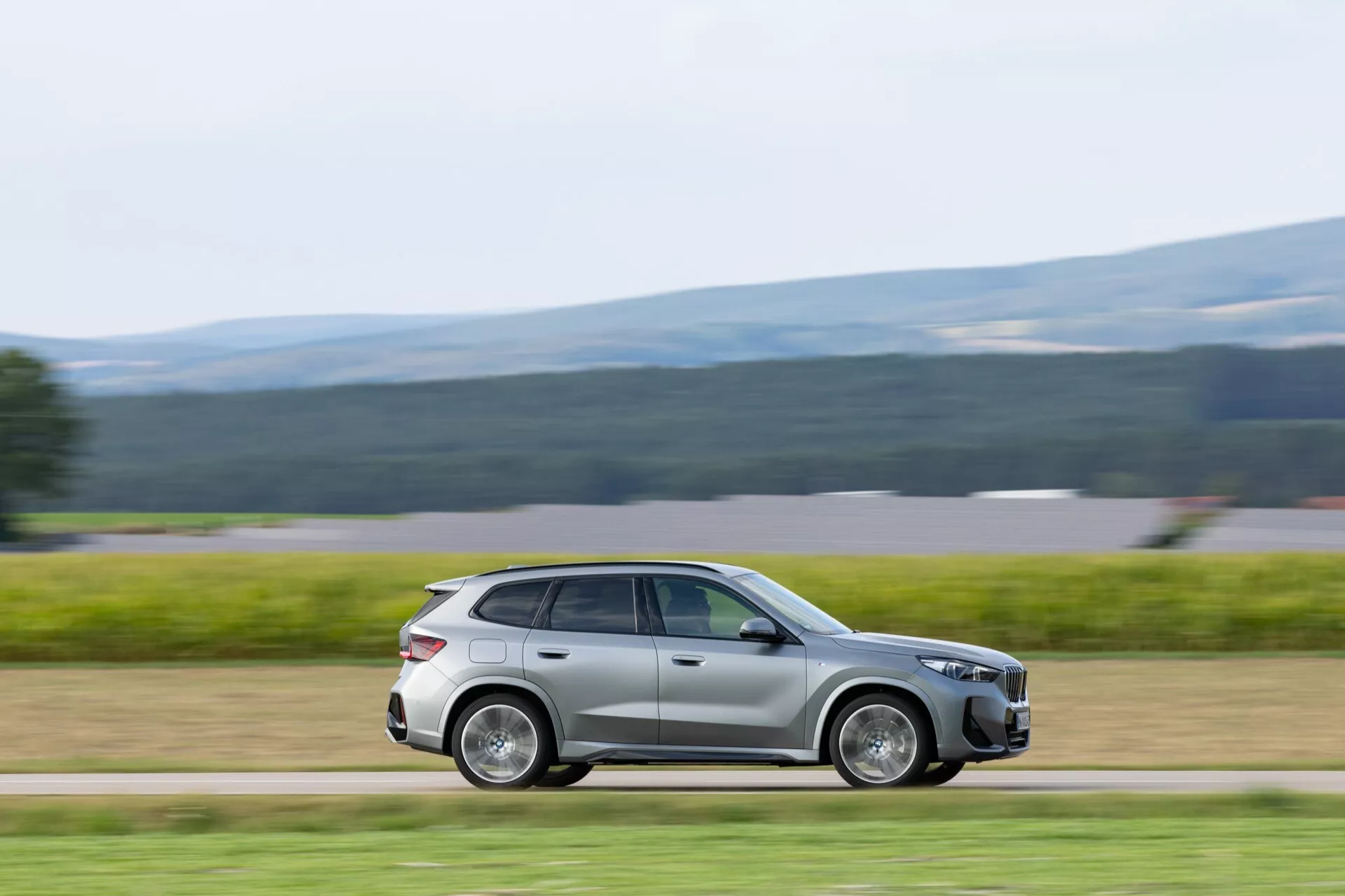 Obraz uchwyca nowoczesny kompaktowy SUV BMW iX1 2022 (U11) z bocznego profilu w dynamicznym, rozmazanym ujęciu, sugerując, że samochód jest w ruchu. Zdjęcie podkreśla elegancki styl auta, wyższy prześwit, design kół oraz aerodynamiczne linie nadwozia, tworząc wrażenie prędkości i podkreślając zaawansowane funkcje i elementy designu w dobrze oświetlonym otoczeniu.