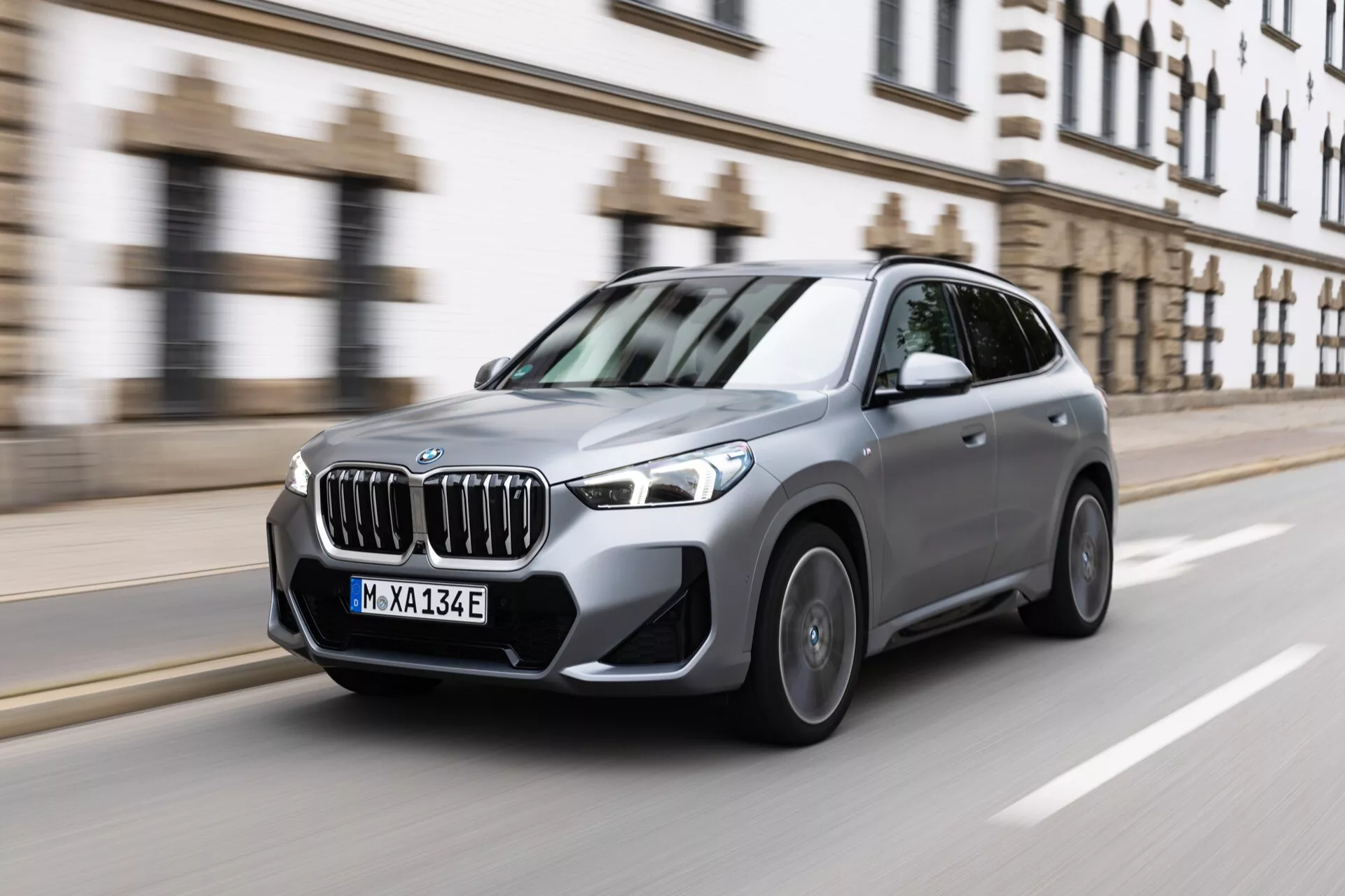 Obraz przedstawia średnie ujęcie SUV BMW iX1 2022 (U11) w ruchu na ulicy miejskiej, uchwycone dynamicznie z trzech czwartych przodu. Pojazd ma wyrazisty grill, smukłe, kątowane reflektory LED, widoczne wloty powietrza, wyprofilowane linie maski, aluminiowe felgi, zdefiniowane linie boczne i zintegrowane kierunkowskazy w lustrach bocznych. Rozmycie w ruchu kół i tła sugeruje umiarkowaną do wysokiej prędkość, ukazując wydajność i zwrotność SUV-a w warunkach miejskich. Obraz sugeruje zaawansowane funkcje technologiczne, takie jak adaptacyjne reflektory LED i systemy wspomagania kierowcy.
