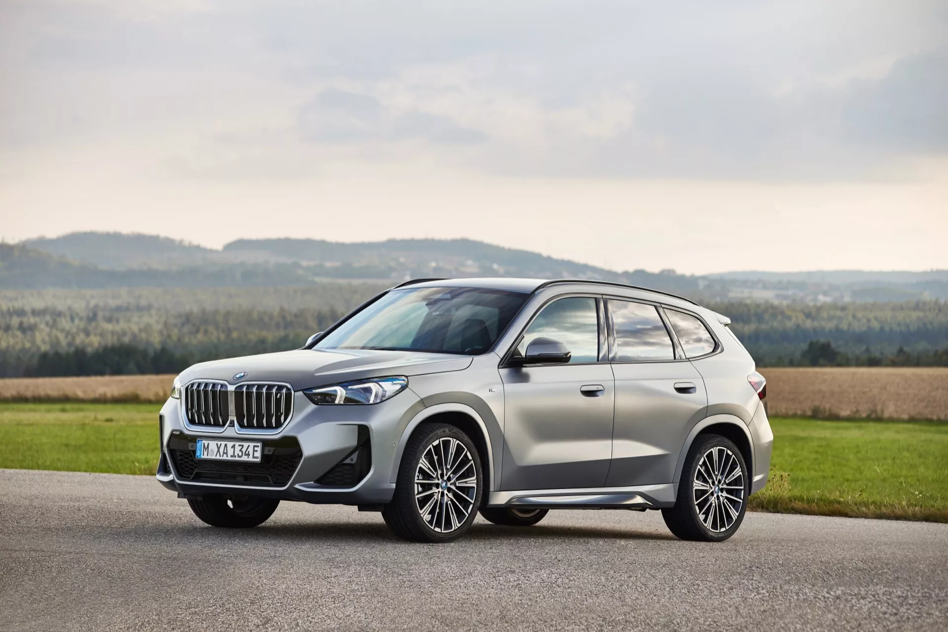 Widok z profilu na SUV BMW iX1 (U11) 2022, prezentujący nowoczesne elementy designu, takie jak wyrazisty przedni grill, reflektory LED, duże felgi aluminiowe i chromowane wykończenia na asfaltowej drodze z malowniczym krajobrazem wiejskim, podkreślone miękkim światłem dziennym.