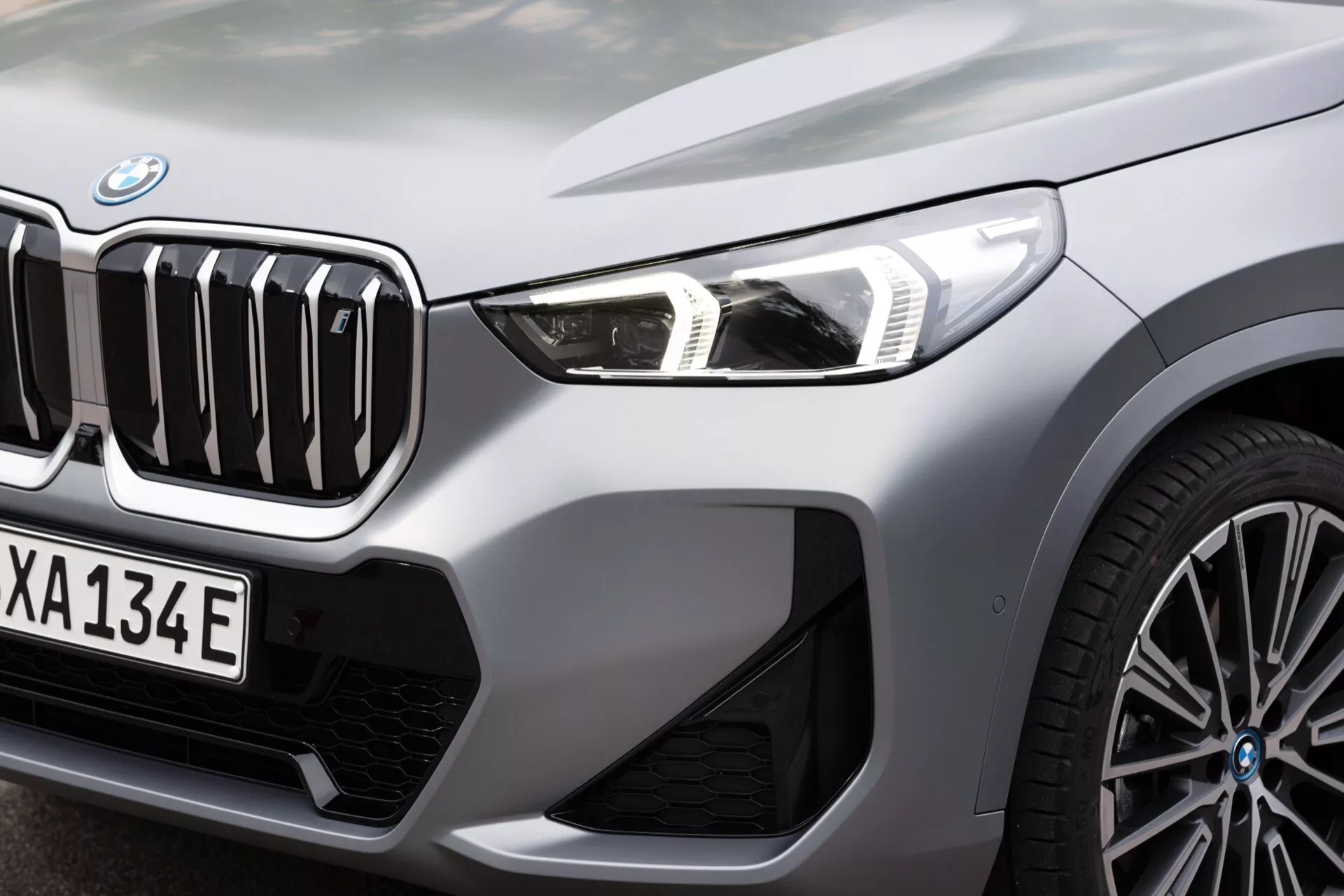 Zbliżenie przedniej, lewej strony BMW iX1 2022 (U11) prezentującej charakterystyczną nerkową osłonę chłodnicy z pionowymi listewkami, smukłe reflektory LED z dziennymi światłami oraz część wieloramiennego aluminiowego koła. Zdjęcie wykonane z niskiego kąta, podkreślające system wlotu powietrza otoczony chromowanymi elementami, a także 'e' na tablicy rejestracyjnej i kratce, wskazujące na model hybrydowy lub w pełni elektryczny. Metaliczne wykończenie podkreśla ekskluzywny i sportowy wygląd pojazdu.