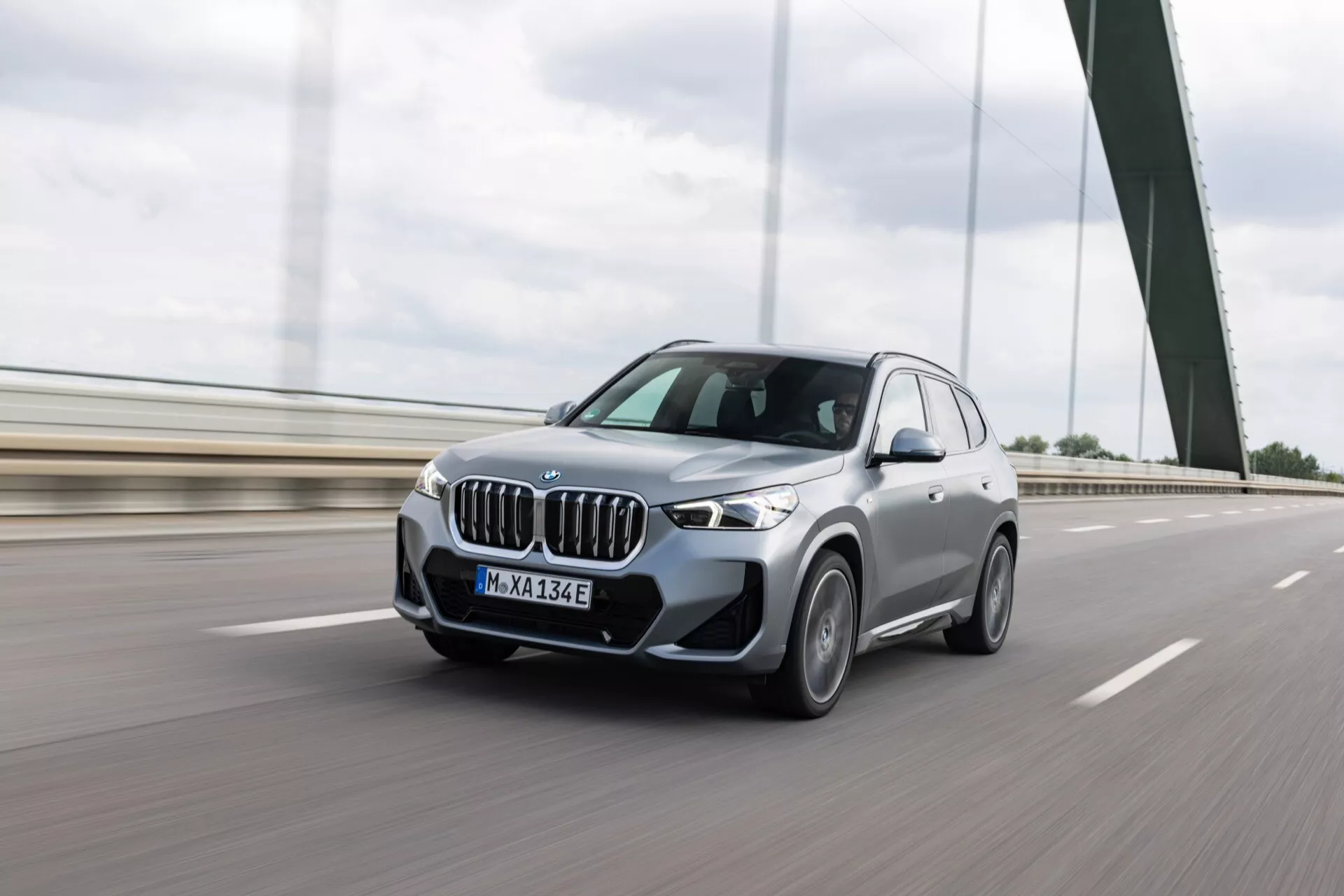 Niski widok z przodu pod kątem trzech czwartych na elektryczny SUV BMW iX3 2022 podróżujący autostradą, ukazujący ikonową nerkową osłonę chłodnicy, reflektory LED, aerodyIsignionamiczny design i rozmycie ruchu na kołach. Zdjęcie robione w ciągu dnia pod pochmurnym niebem.