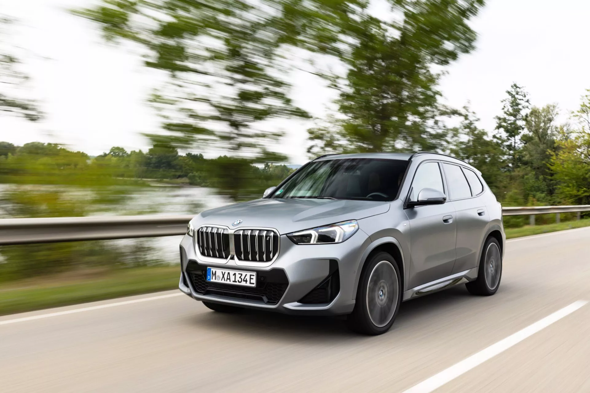 BMW iX1 2022 (U11) nowoczesny SUV jadący prawą stroną dwupasmowej drogi, zbliżający się do kamery. Pojazd jest uchwycony w ruchu z rozmytym tłem, co wskazuje na prędkość. Widok od przedniego trzech czwartych pokazuje charakterystyczną nerkę, kanciaste reflektory LED, wieloramienne felgi aluminiowe i metaliczny lakier jasnoszary, z widoczną odznaką 'e', sugerującą model zelektryfikowany.