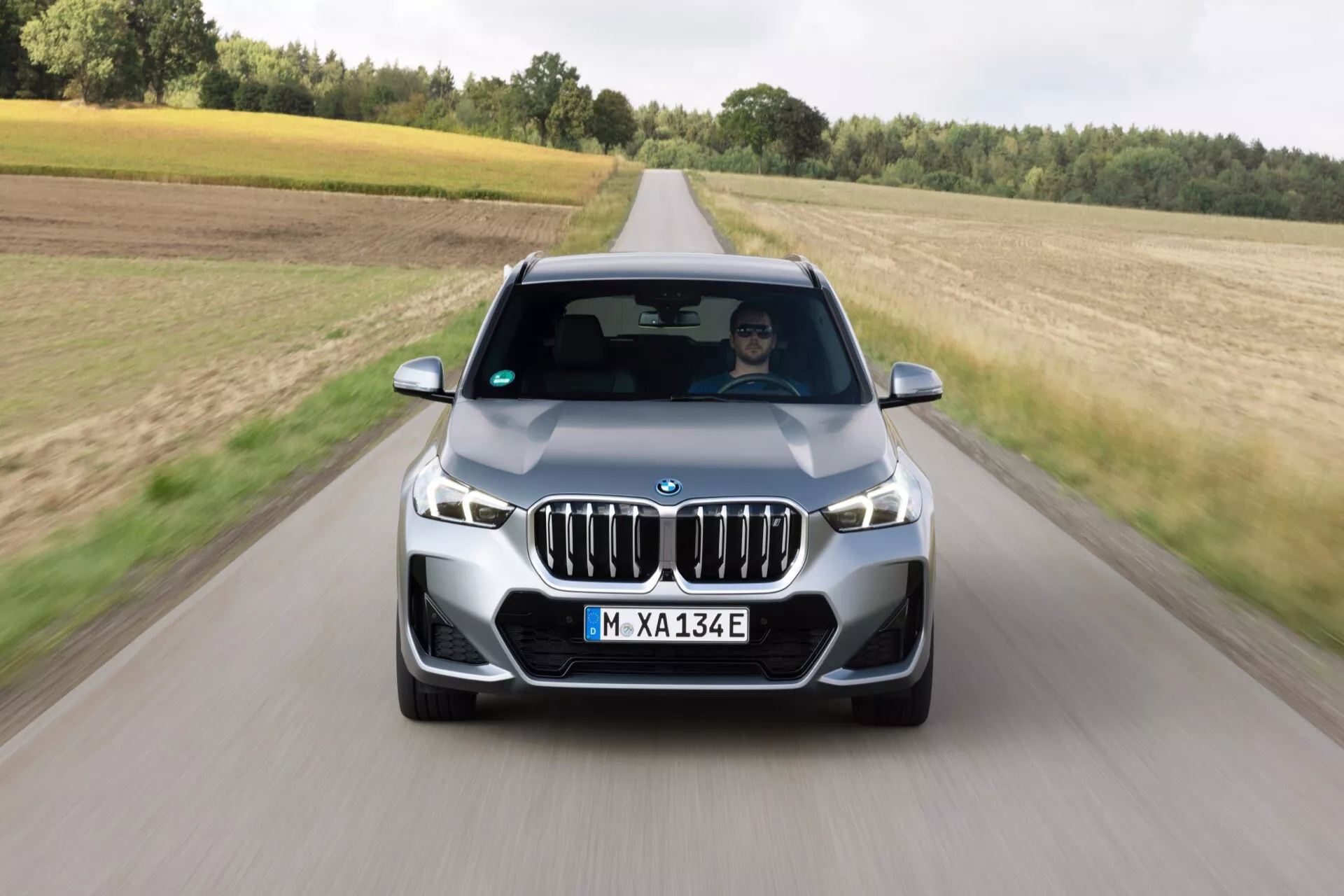 Widok z przedniego kąta na BMW ix1 2022 (U11) w ruchu na utwardzonej drodze, ukazujący charakterystyczny grill w kształcie nerek pojazdu, pełny widok z przodu i stylowy design. Ujęcie wykonane z nieznacznie podwyższonej pozycji, podkreślające wąskie i cofnięte reflektory, wyrazisty przedni grill z pionowymi listwami oraz tablicę rejestracyjną z oznaczeniem 'M-XA 134E'. Dynamiczny wygląd samochodu został uchwycony w półwiejskim krajobrazie z polami i drzewami pod pochmurnym niebem. Widoczna jest osoba na fotelu kierowcy, co podkreśla, że samochód jest w ruchu.