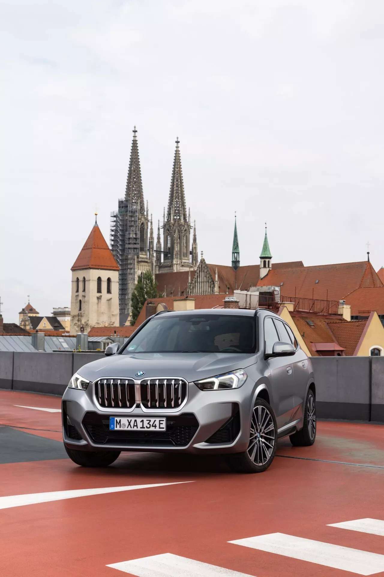 Widok z przodu i z boku na SUV BMW iX1 (U11) z 2022 roku zaparkowany na świeżym powietrzu, podkreślający dużą kratkę chłodnicy w kształcie nerki i niebieskie akcenty wskazujące na jego elektryczny lub hybrydowy plug-in charakter. Pojazd wyposażony jest w reflektory LED, alufelgi i czujniki parkowania. Znajduje się na tle zabytkowej europejskiej architektury, w tym katedry z wieżami, zaparkowany na górnym poziomie parkingu z czerwoną strefą oznakowania.