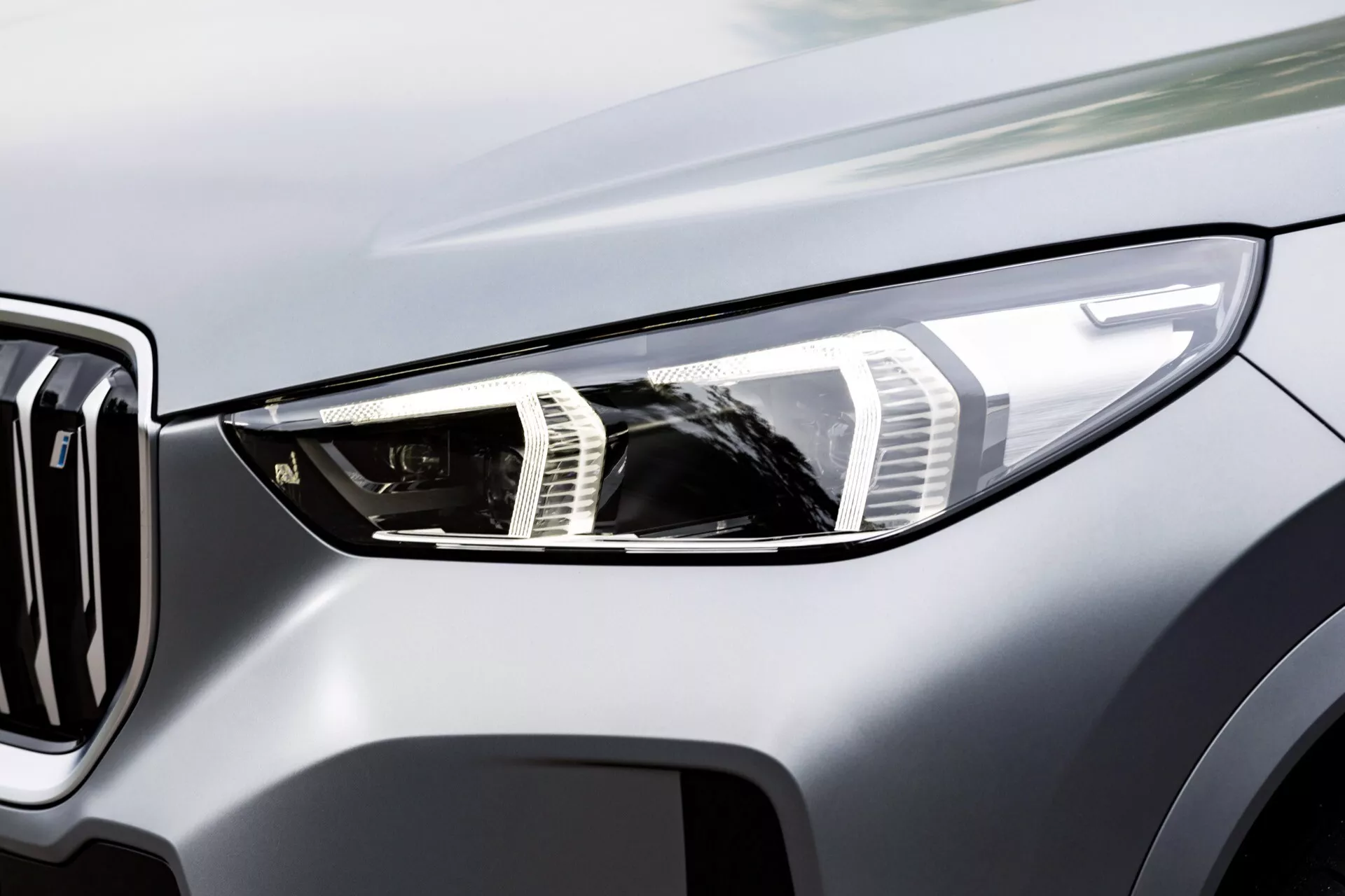 Zbliżenie na obszar reflektorów samochodu BMW iX1 2022 (U11) z boku, skupiając się na lewym przodzie. Obraz ukazuje lewy reflektor, część grilla i fragment maski z złożonymi diodami LED do jazdy dziennej i zaawansowanym wzorem grilla w metalicznym wykończeniu.