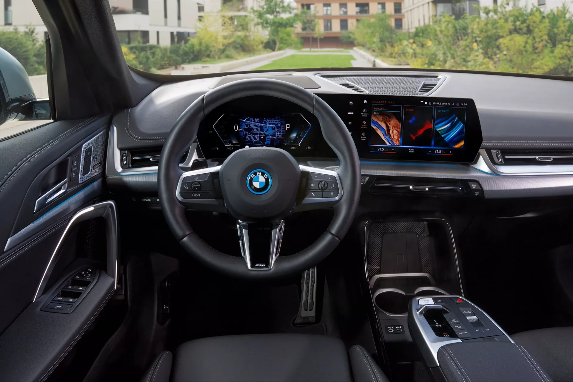 Widok wnętrza BMW iX1 2022 (U11) z tylnej części kabiny, przedstawiający nowoczesny kokpit z kierownicą trójramienną z logo BMW i przyciskami sterującymi, niebieski cyfrowy zestaw wskaźników wyświetlający prędkość i obroty, centralny ekran dotykowy systemu multimedialnego, minimalistyczne fizyczne przyciski, konsola centralna z dźwignią zmiany biegów i pokrętłem iDrive, luksusowe materiały, takie jak skóra i metalowe lub błyszczące wykończenia na desce rozdzielczej i panelach drzwi, oświetlenie nastrojowe na panelach drzwi oraz częściowo widoczne skórzane przednie siedzenia w kolorze czarnym.