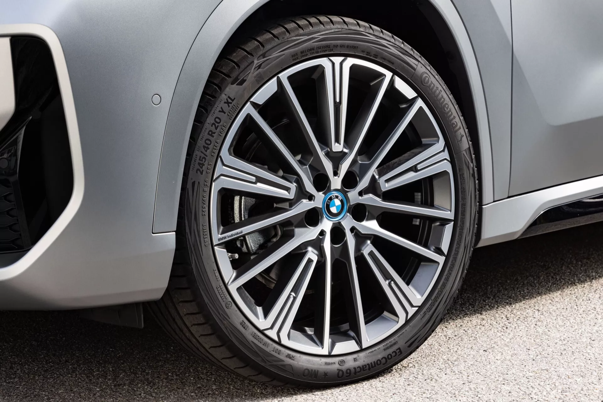 Zbliżenie przedniego prawego koła BMW iX1 2022 (U11), sfotografowane z poziomu ziemi z nieco przekątnej perspektywy. Felga wieloramienna z mieszanym wykończeniem polerowanym i ciemnym podkreśla luksusowy, sportowy trim. Środkowa nakładka zawiera logo BMW. Opona Bridgestone DriveGuard typu run-flat pokazuje widoczny wzór bieżnika i napisy na ścianie bocznej. W tle widoczna metaliczna karoseria z zakrzywionym przednim błotnikiem.