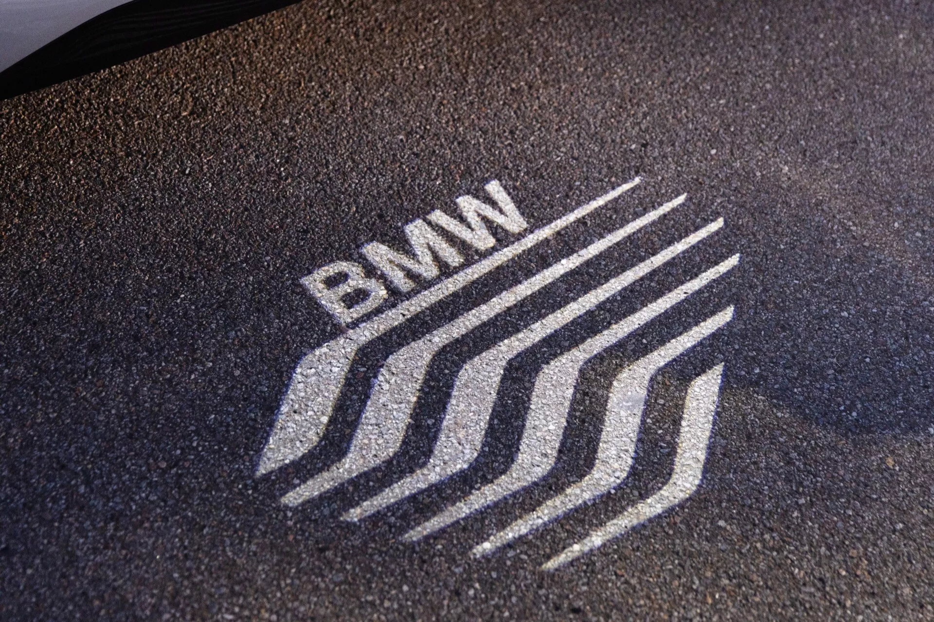 Zbliżenie stylizowanej projekcji emblematu BMW na teksturowanej powierzchni, prawdopodobnie asfalcie. Oświetlone logo ma dynamiczne linie zamiast tradycyjnego okrągłego wzoru. Ten efekt świetlny prawdopodobnie pochodzi z zewnętrznego oświetlenia pojazdu BMW, takiego jak światło kałuży, podkreślając innowacyjne elementy oświetlenia ambientowego i brandingu.