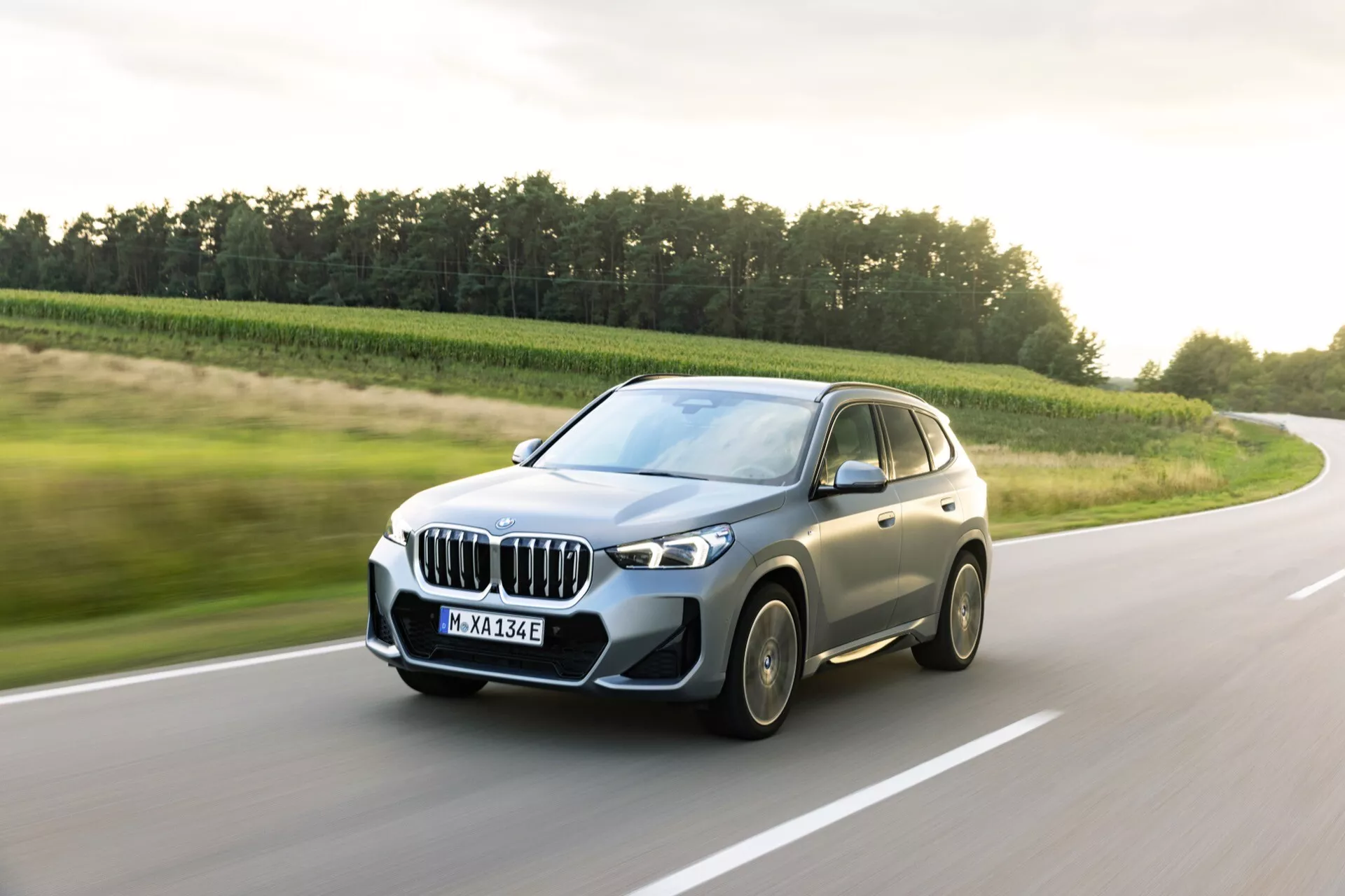 BMW iX1 2022 (U11) w ruchu, uchwycony pod kątem ćwiartki przedniej, pokazujący charakterystyczną nerkową kratkę, reflektory LED z DRL, czujniki na przednim zderzaku, felgi aluminiowe i lusterka boczne z zintegrowanymi kierunkowskazami. Aerodynamiczna karoseria ma ostre linie, możliwy panoramiczny dach i wyraźny widok do wnętrza przez przednie boczne okno. Rozmazane koła wskazują na ruch, sugerując pewnie prowadzenie wspomagane zaawansowaną technologią zawieszenia. Odznaka 'eDrive' na przednim błotniku sugeruje napęd hybrydowy lub w pełni elektryczny. Fotografowane w świetle dziennym, z dynamicznie niższego kąta na utwardzonej wiejskiej drodze.