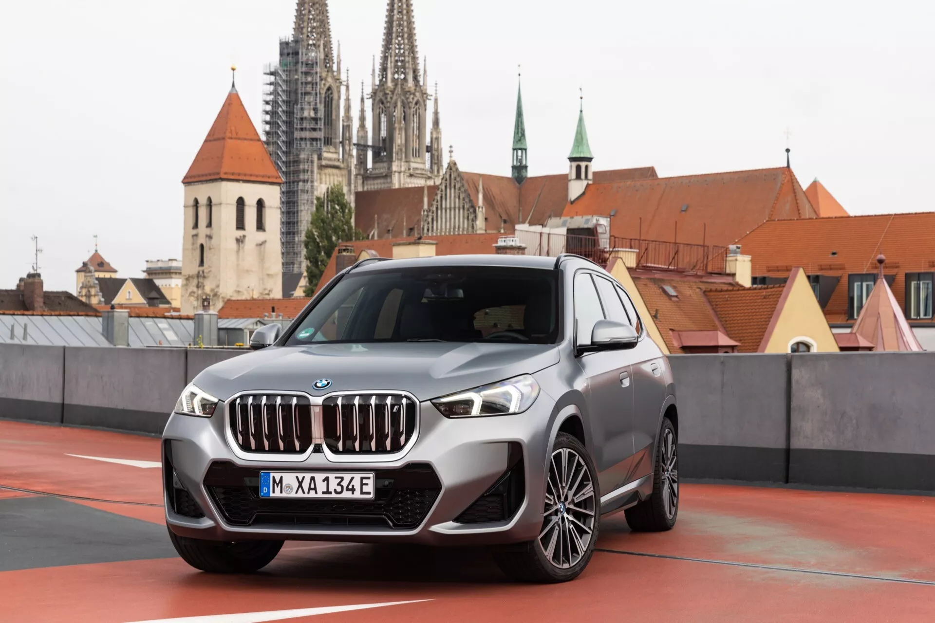 Obraz przedniego kwartału nowoczesnego SUV-a BMW iX1 2022 (U11) z charakterystycznym grillem nerkowym, eleganckimi reflektorami LED i skomplikowanymi felgami aluminiowymi. Pojazd jest zaparkowany na dachu z widoczną starą europejską architekturą, w tym wyraźną gotycką iglicą katedry w tle, sugerującą europejskie otoczenie miejskie.