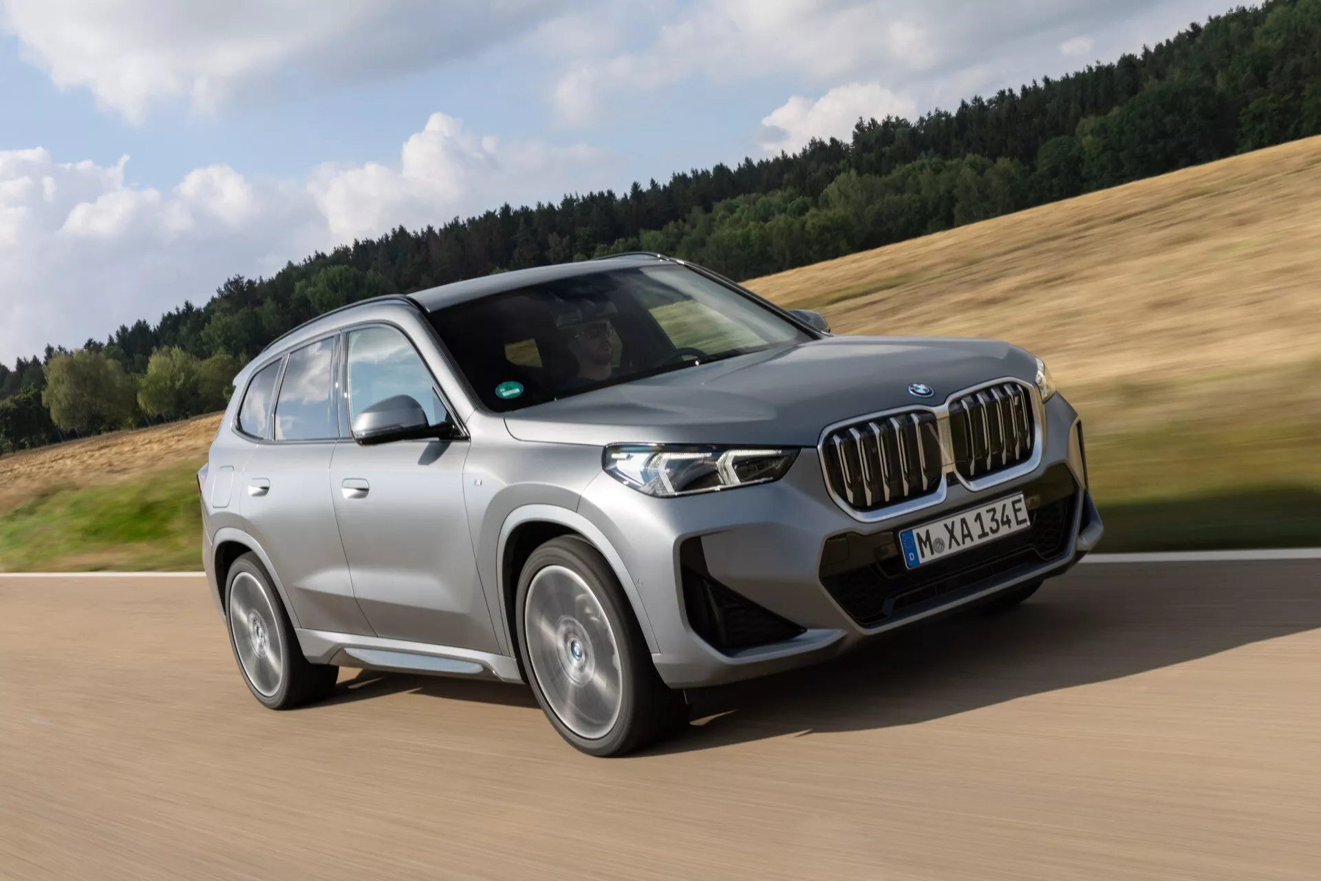 Dynamiczne zdjęcie SUV-a BMW iX1 2022 (U11) w ruchu, zrobione z niskiego kąta od przodu po lewej stronie, aby pokazać przednią fasadę, profil strony kierowcy i nowoczesne elementy projektowe. Charakterystyczny grill nerkowy, reflektory LED, wieloszprychowe felgi aluminiowe, aerodynamiczny zderzak i eleganckie lusterka sugerują zaawansowane osiągi i najnowocześniejszą technologię, w tym możliwe zdolności modelu hybrydowego lub elektrycznego. Rozmazane tło podkreśla prędkość i dynamikę jazdy.