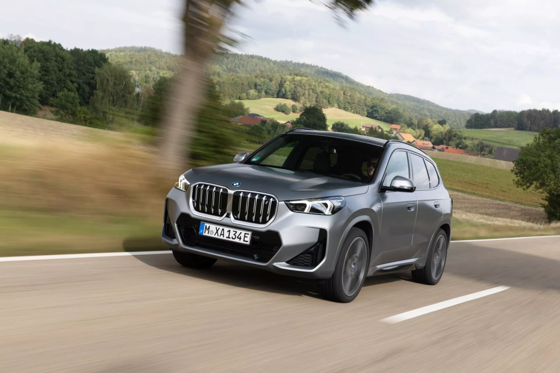 Ruchomy BMW iX1 2022 (U11) SUV, z trzy-czwiartkowego kąta przedniego, ukazującego lewą przednią stronę, charakterystyczna pionowa kratka, ostre światła LED, przedni zderzak z czujnikami, aerodynamiczny kształt, duże lusterka z wbudowanymi kierunkowskazami, wieloramienne alufelgi, w ruchu rozmytym na tle zieleni i błękitów