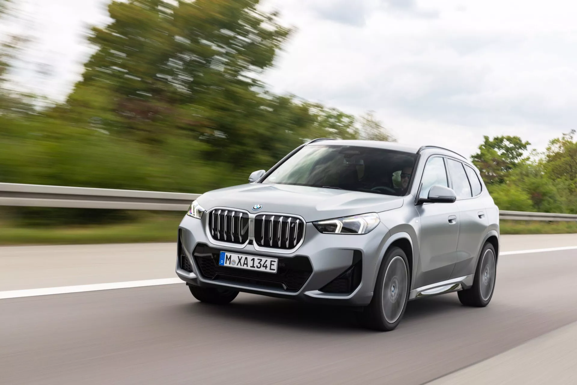 Obraz BMW iX1 2022 (U11) w ruchu na autostradzie, uchwycony z lekkim rozmyciem ruchu wokół kół i tła. Widok przedniej ćwiartki pokazujący przedni grill, reflektory, przedni zderzak, koła, lusterka boczne i okna. Nowoczesne reflektory LED, skomplikowany wzór grilla z czujnikami do zaawansowanych systemów asystujących kierowcy, przedni dolny zderzak z czujnikami parkowania, przednia kamera w grillu do funkcji autonomicznej jazdy, aerodynamiczne koła, lusterka z systemem monitorowania martwego pola i 'E' na tablicy rejestracyjnej wskazującej na technologię elektryczną lub hybrydową. Pojazd jest umieszczony w lewym pasie, prezentując elegancki i sportowy design luksusowego SUV-a z zaawansowaną technologią samochodową.