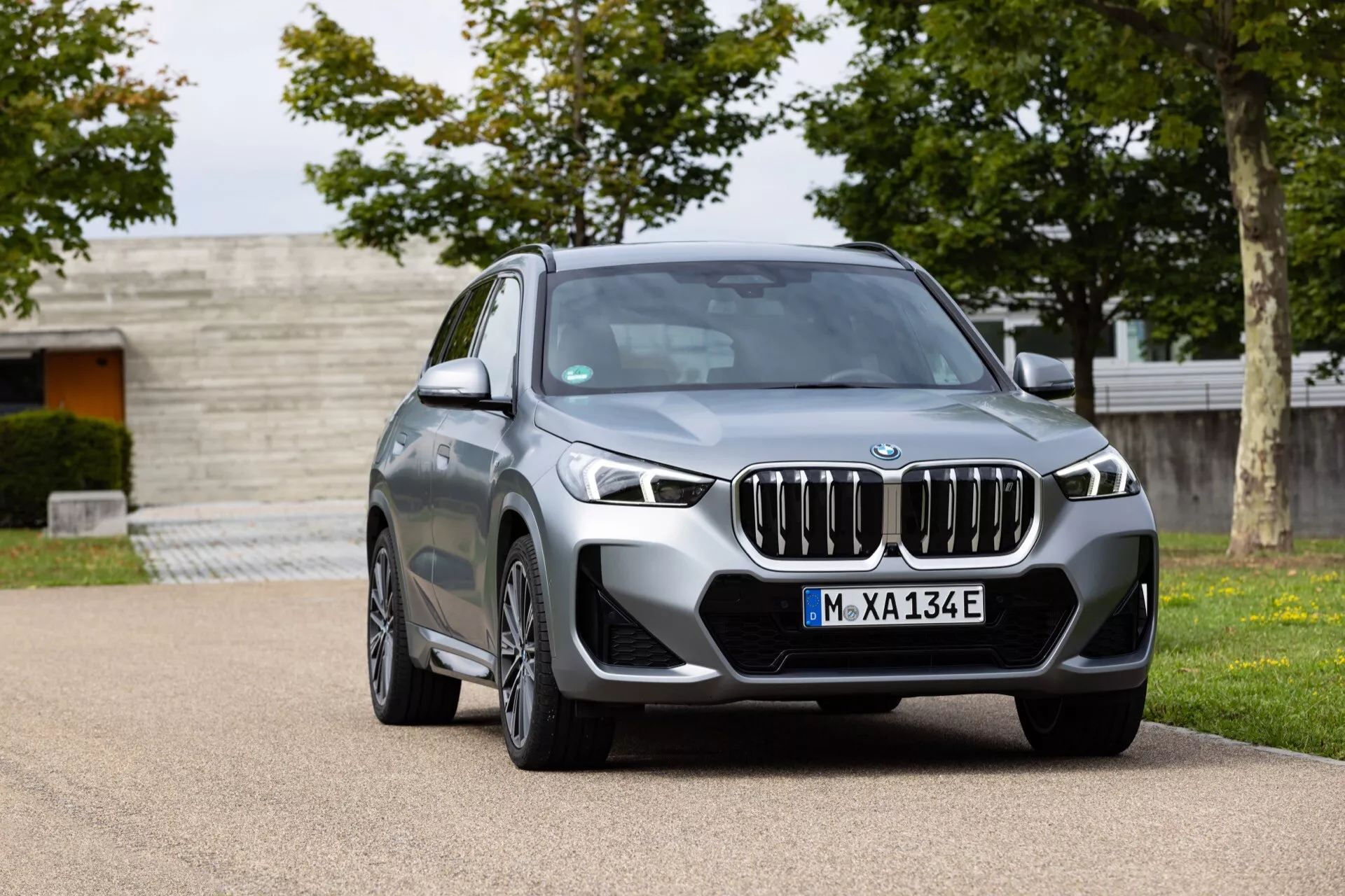 Obraz nowoczesnego luksusowego SUV-a BMW iX1 2022 (U11) zaparkowanego na kamienistej nawierzchni w otoczeniu rezydencjalnym lub parkowym. Pojazd jest ustawiony pod kątem trzech czwartych frontalnie, prezentując swoją szeroką nerkową osłonę chłodnicy, eleganckie reflektory LED z dziennymi światłami, duże wloty powietrza, wyraźne linie maski z centralnym emblematem BMW, aerodynamicznie zaprojektowane lusterka z wbudowanymi kierunkowskazami, wieloramienne felgi aluminiowe, okna z chromowanymi akcentami, szyberdach i metaliczny szary lakier. Tablica rejestracyjna 'M-XA134E' sugeruje, że może być elektrycznym albo hybrydowym modelem typu plug-in.