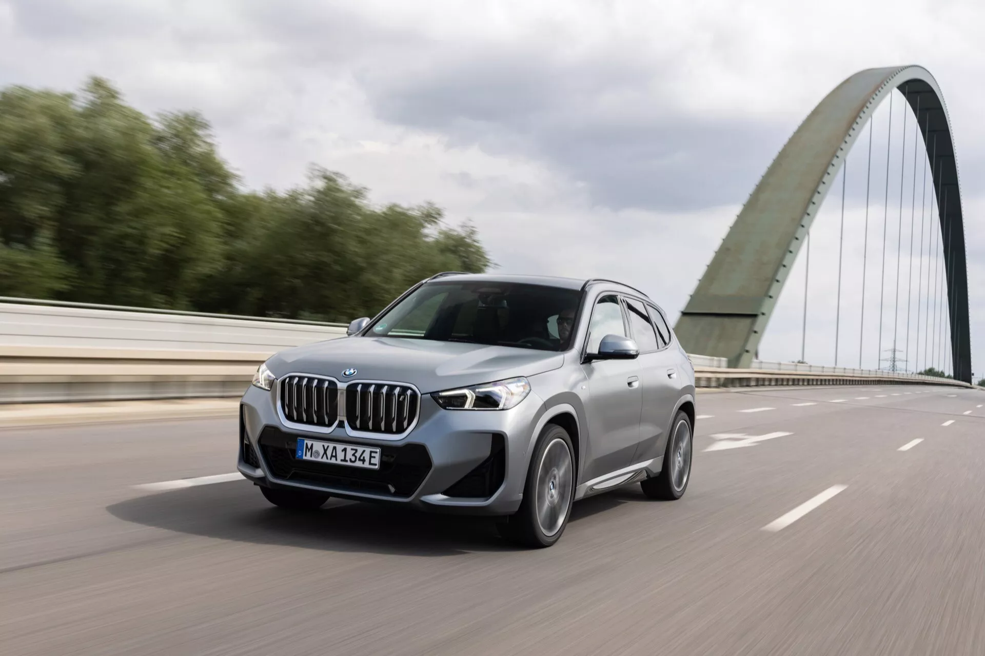 Obraz przedstawia srebrne BMW iX1 2022 (U11) SUV w ruchu na wielopasmowej autostradzie. Uchwycone pod kątem trzech czwartych z przodu, wyróżnia się dużą kratką nerkowatą, eleganckimi reflektorami LED, wieloszprychowymi aluminiowymi felgami i robustnym nadwoziem. Samochód, wyposażony w lusterka boczne z wskaźnikami kierunku i odznaki wskazujące na model hybrydowy lub elektryczny, emanuje luksusem i nowoczesnością. W tle znajduje się most, wzmacniając temat dynamicznej jazdy na infrastrukturze publicznej.