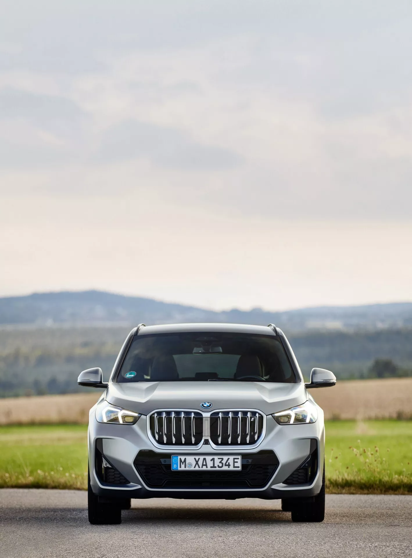 Widok z przodu BMW iX1 2022 (U11), eksponujący charakterystyczny grill nerkowy, smukłe adaptacyjne reflektory LED i rzeźbione linie maski. Elektryczny lub hybrydowy pojazd stoi na drodze z trawiastym poboczem, z rozmytym wiejskim tłem podkreślającym jego wyrafinowany design i zaawansowaną technologię oświetleniową oraz aerodynamiczną.